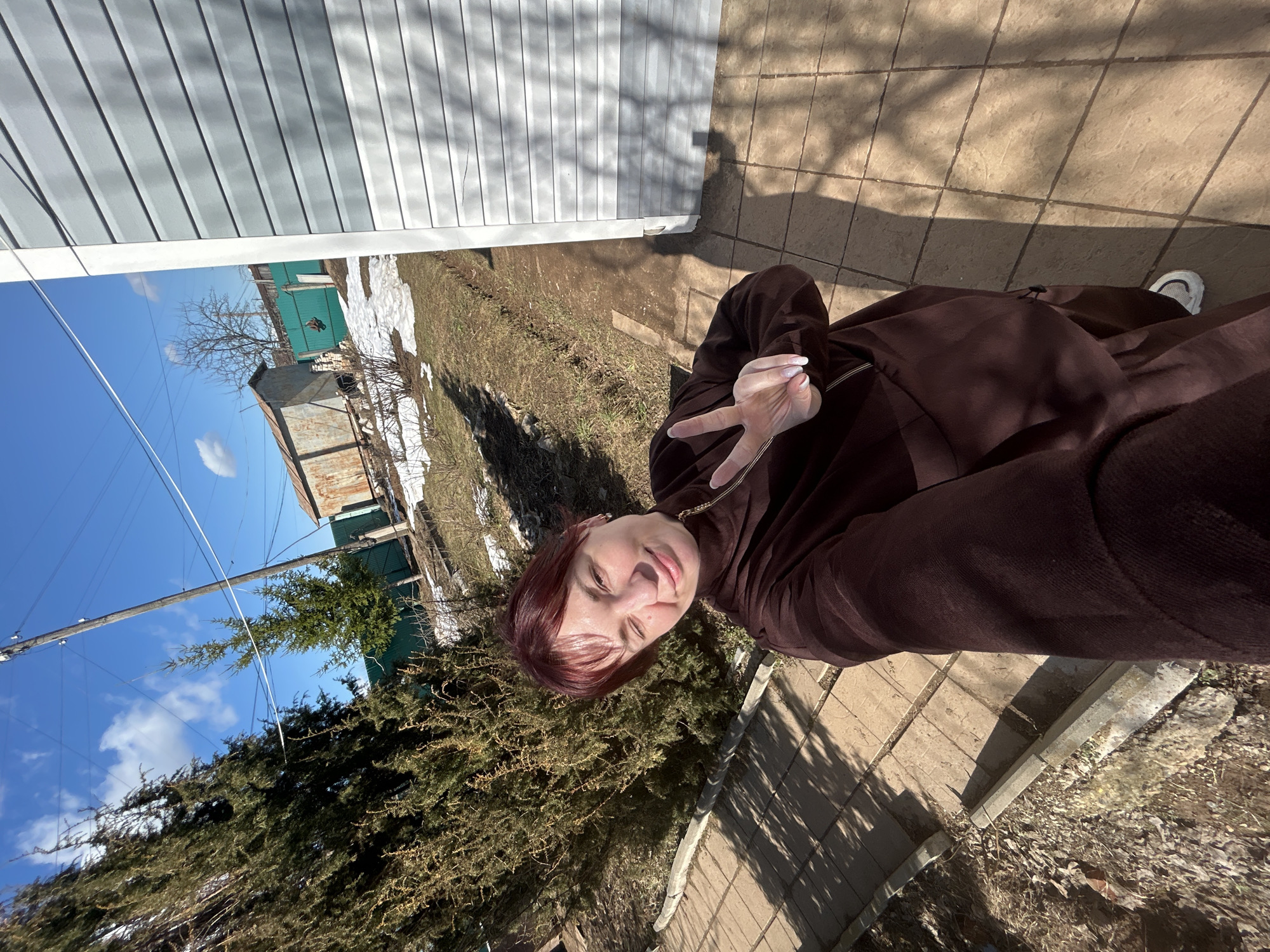 A selfie of a young, fair-skinned woman with short, auburn hair and a brown coat stands outside on a sunny day. She has a peace sign up with her hand. She stands on a stone-paved walkway. Behind her is a yard with patches of grass and snow. In the background is a light blue house and a corrugated metal structure, possibly in Фоки, Russia, based on the query. A wooden electrical pole and a blue sky are also visible.