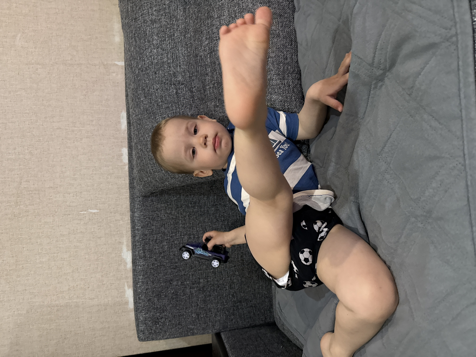 This media file captures a young child lying on a gray upholstered couch in what appears to be an indoor setting, likely a home in Rusanovo, Russia. The child, a toddler with light hair, is wearing a blue and white striped t-shirt and black shorts or a diaper cover adorned with white soccer ball patterns. One of the child's legs is raised high in the air, showcasing the sole of their foot, while the other leg is bent. The child's gaze is directed towards the camera, with a neutral to slightly curious expression.
In their left hand, the child holds a small, dark blue or purple toy car resembling a jeep or truck. The couch itself is covered with a gray, possibly quilted, fabric on one side. To the left of the child and couch, a light beige or off-white textured wallpaper is visible, showing some subtle imperfections or marks. The overall lighting suggests it is daytime. On the child's t-shirt, faint text reading "Sea You" or similar is visible.
