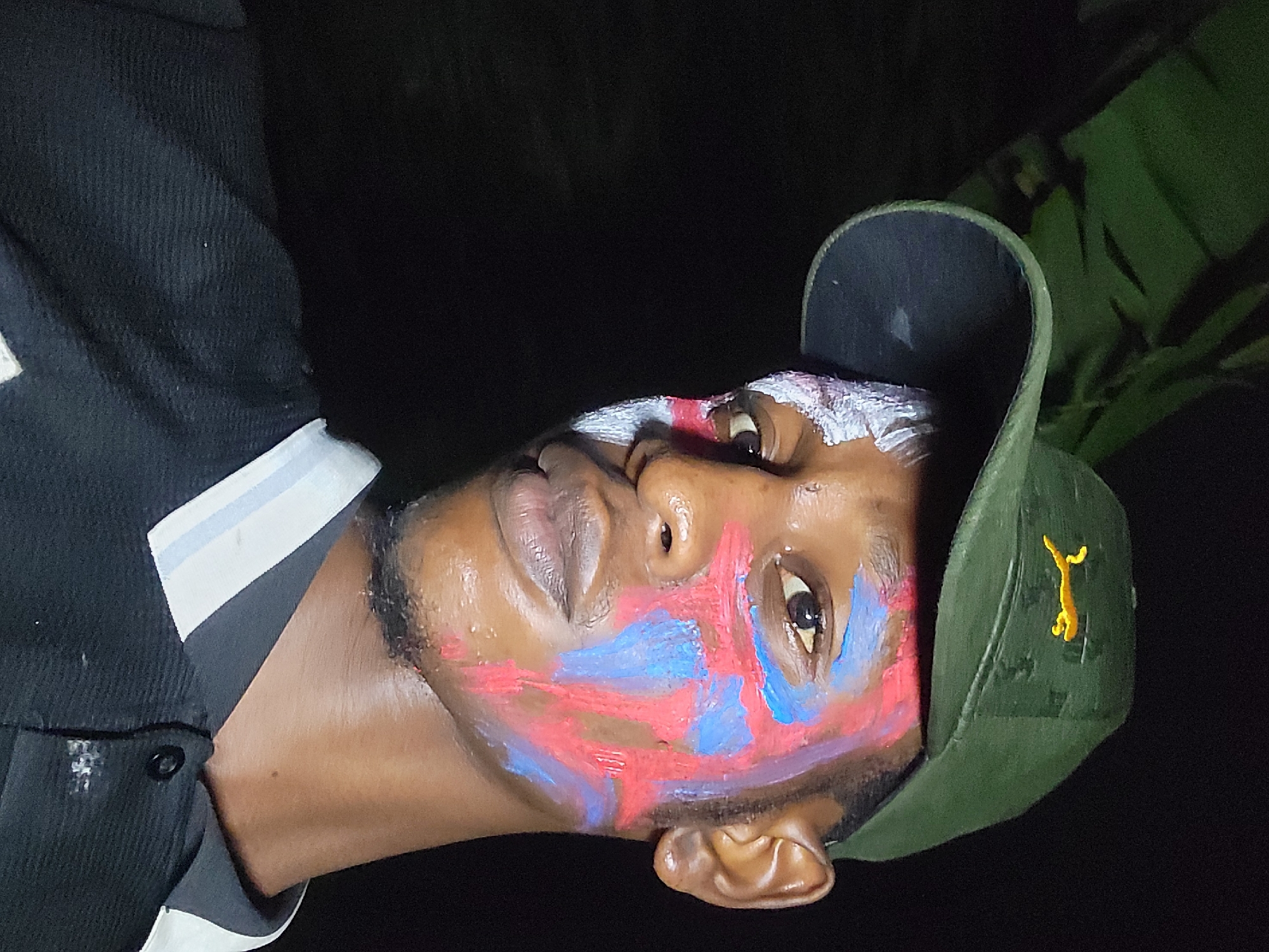 A close-up, vertically oriented, low-angle photograph depicts a Black man with face paint in Nkpor, Nigeria. The man wears an olive green cap with a yellow logo resembling a stylized animal on its right side. His face is adorned with broad strokes of red and blue paint across his cheeks and forehead, with white paint near the bridge of his nose and eyebrows. The man’s lips are slightly parted. He is wearing a black jacket with white and light blue trim. The background is predominantly dark, with a hint of green foliage visible in the upper right corner.