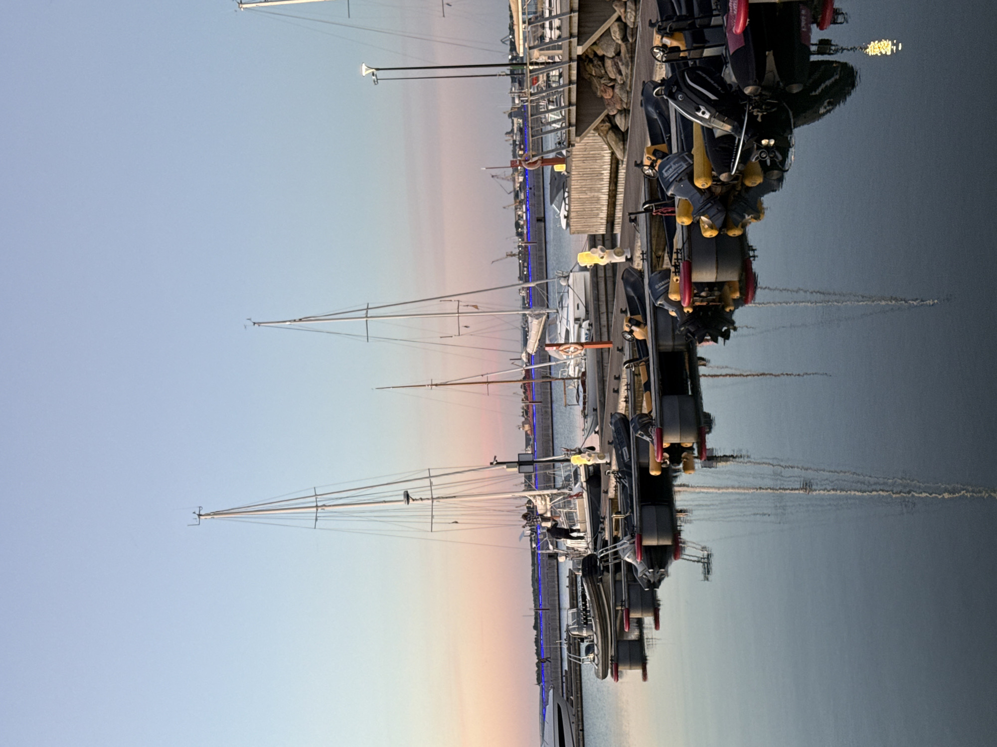 The image captures a serene harbor scene at dusk in Tallinn, Estonia, likely the Pirita Marina. The sky transitions from a pale blue to soft pink and orange hues near the horizon, reflecting on the calm water. Several sailboats and smaller inflatable boats are docked. One person is visible on the deck of a sailboat, seemingly engaged in some activity. The marina is equipped with wooden docks and mooring facilities. In the background, a distant cityscape with industrial cranes and structures can be observed. The overall atmosphere is tranquil, illuminated by the fading daylight and a few artificial lights.