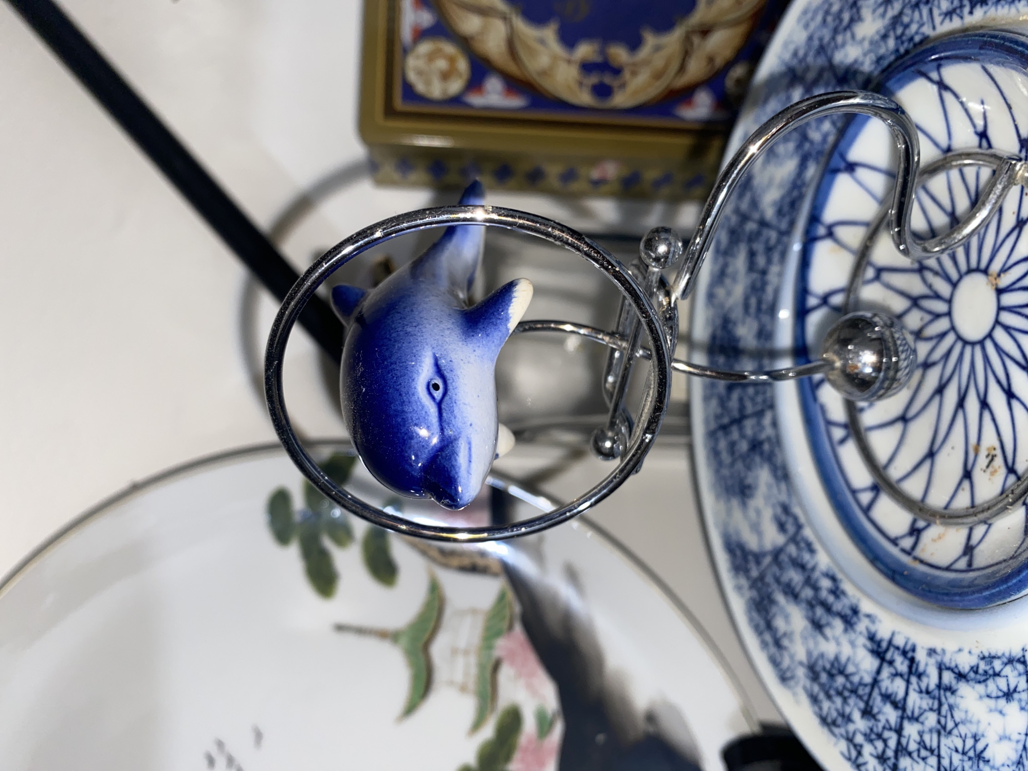 The image shows a close-up of a small, ceramic blue and white dolphin figurine held within a silver, circular stand. The stand also holds several plates. One plate has a blue and white floral pattern. Another plate features a white background with green and pink floral and branch designs. Above, a box with gold accents and blue designs is visible. A black rod extends diagonally across the upper left corner of the frame. The location is not discernible from the image.