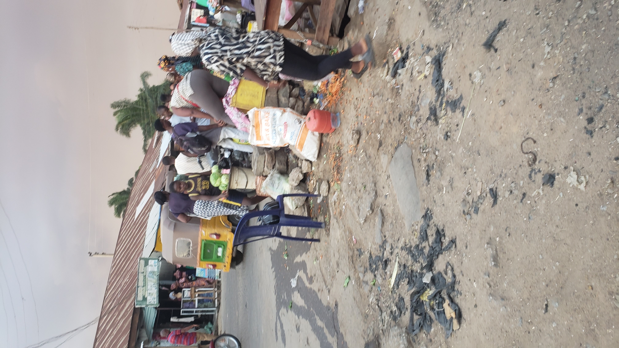 The image depicts a scene in Narayi, Nigeria, likely a market area, due to the presence of multiple people gathered near a structure with a corrugated metal roof.
People: A group of people is seen interacting around the building. Some are standing and appear to be engaged in commerce or conversation. Clothing styles indicate a casual, everyday setting.
Objects: There are bags, containers, and other items suggesting goods being sold or transported. A blue plastic chair is also visible near the building. A yellow tricycle is visible behind people.
Environment: The ground is largely bare and littered with debris. The presence of overhead power lines hints at an urban or semi-urban location. The overall scene suggests an active but possibly unkempt environment.