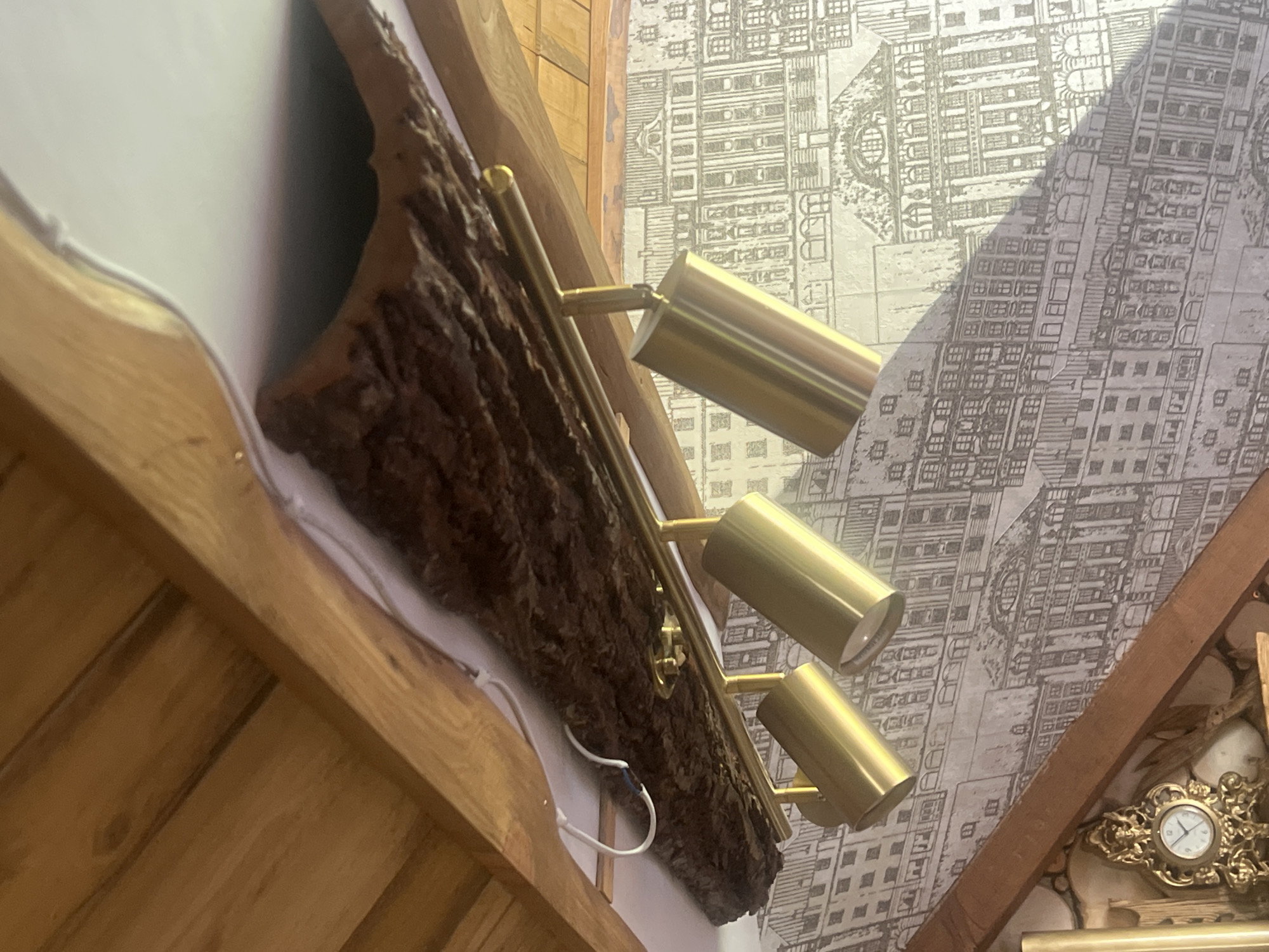 The image depicts an indoor scene featuring a track lighting fixture with three cylindrical, gold-toned spotlights mounted on a golden rail. This rail is attached to a decorative element that appears to be a piece of rough bark or a natural wood beam, which is in turn affixed to a wooden ceiling structure. The background consists of wallpaper with a grey-scale pattern of architectural drawings of buildings. In the lower left portion of the image, a decorative golden clock with intricate scrollwork is partially visible, along with some wooden logs and what appears to be other decorative items. The scene is illuminated by artificial light, casting shadows. There are no people or animals visible in the image. The location is identified as Norviliškės, Lithuania.