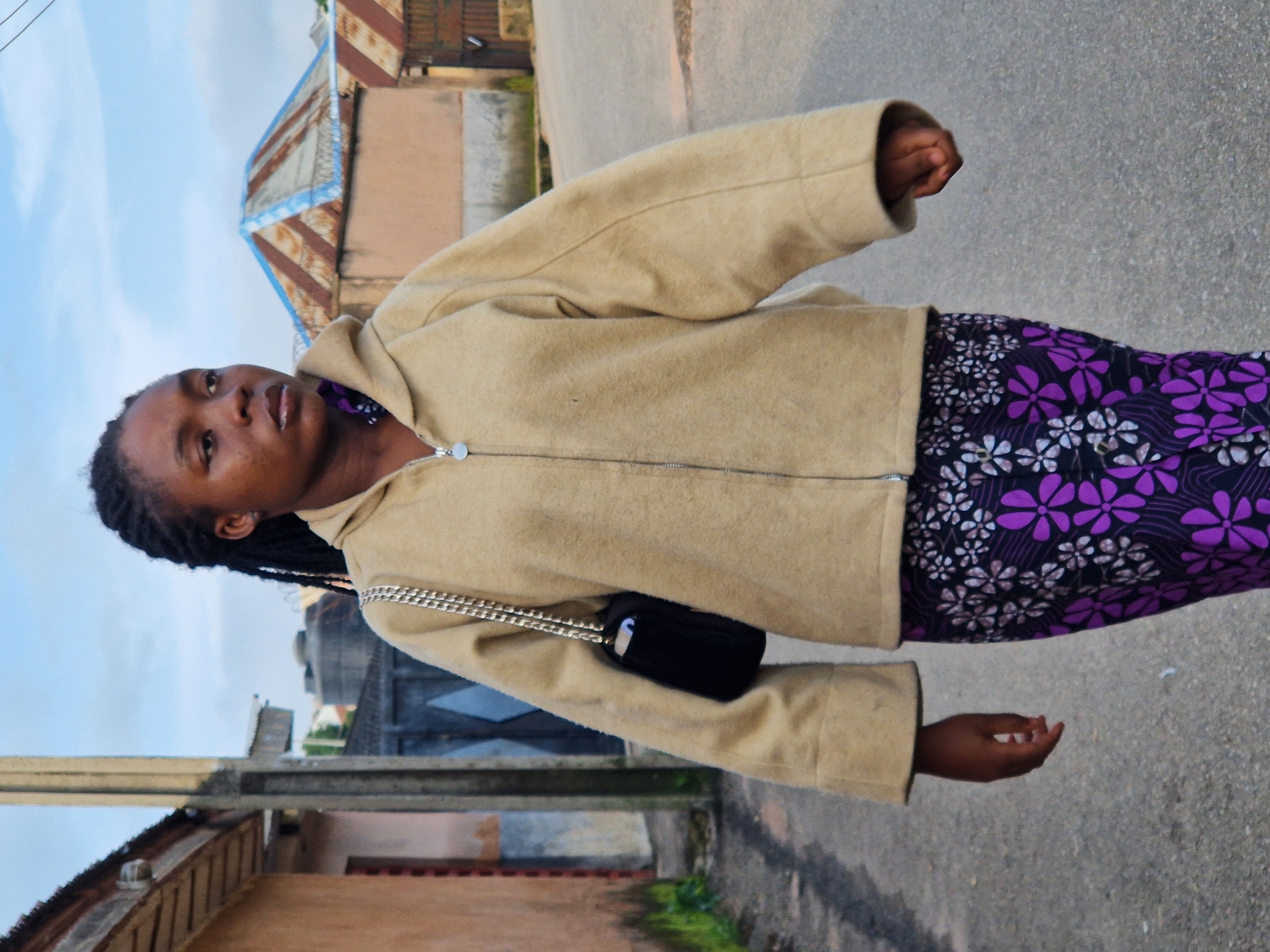 A young Black female with dark braided hair walks on a paved road in Kabong, Nigeria. She is wearing a tan zip-up hooded jacket over a purple and white floral print skirt. A black purse with a silver chain strap is slung over her left shoulder. The background consists of buildings with corrugated metal roofs and a cloudy sky.