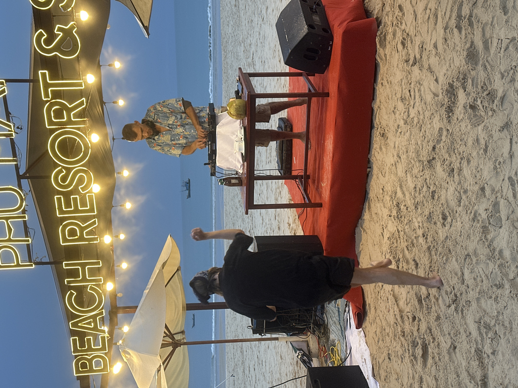 The image depicts an outdoor event on a beach in Phu Thuy Ward, Vietnam. A man in a patterned shirt is DJing at a setup consisting of a table with a DJ console, speakers, and a coconut on top. Another person, wearing a dark shirt and bandana, is dancing near the DJ booth on the sandy beach. A sign that reads "Phu Ly Beach Resort &" is illuminated with lights in the background along with beach umbrellas. The ocean is visible in the distance.