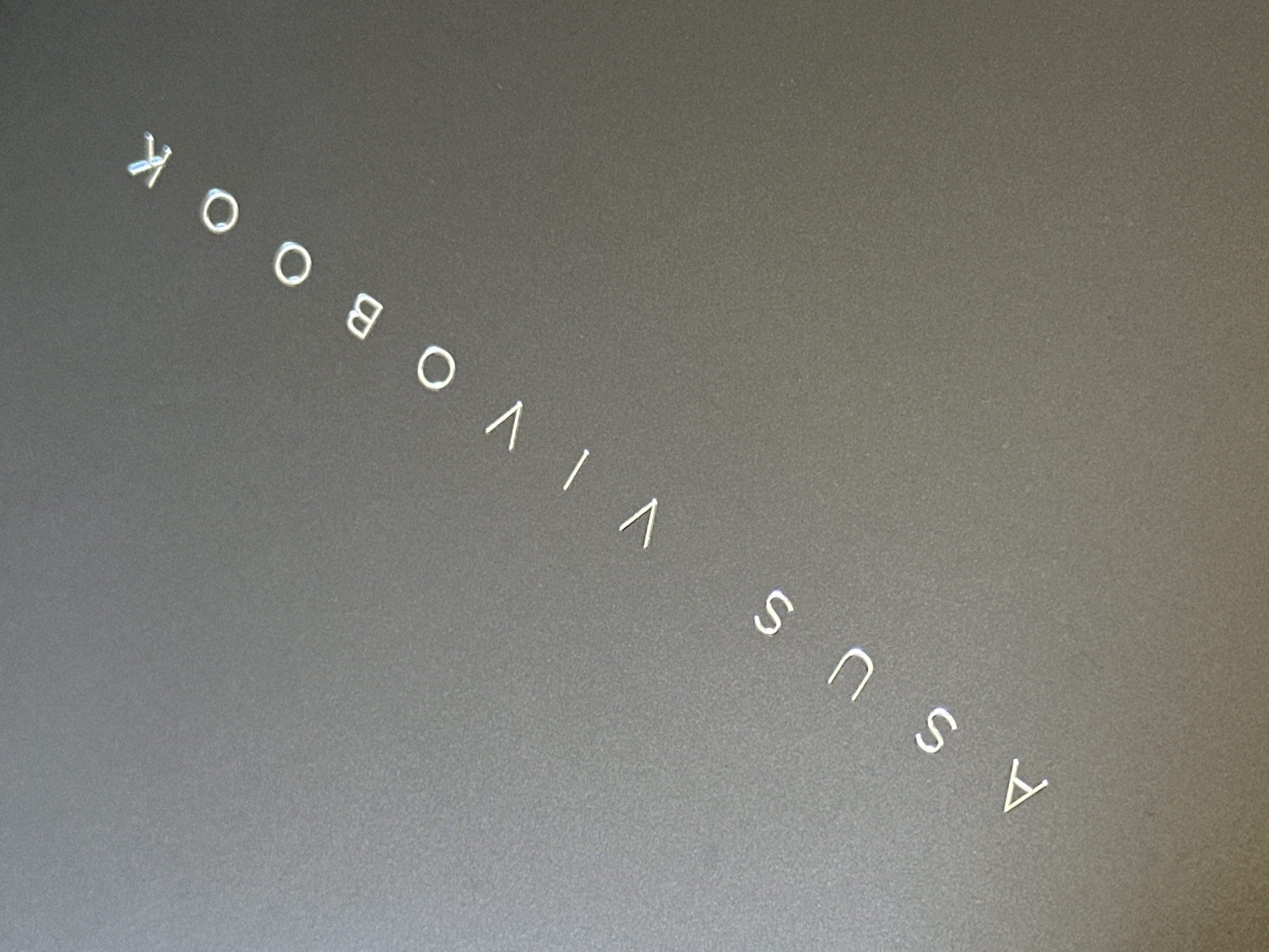 The image shows a close-up of a grey surface with the words "ASUS VIVOOBOOK" written in silver, thin, sans-serif font, diagonally from the bottom left to the top right. The letters are spaced out and appear to be embossed or etched into the surface. The surface itself has a subtle texture, possibly brushed metal or a matte finish. There are no people or other objects visible in the image. The lighting is soft, creating a slight sheen on the metallic lettering. There is no information in the image that can be used to determine its location in Uzhhorod, Ukraine.