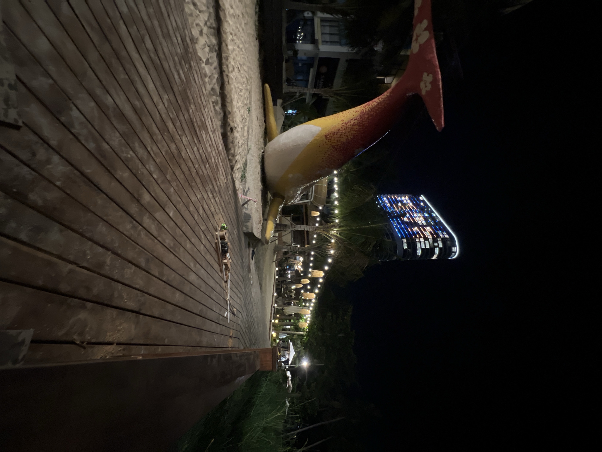 The image shows a nighttime scene along a sandy beach promenade in Nha Trang, Vietnam. A large, colorful sculpture of a whale is positioned on the sand, with its tail fin prominently visible in the foreground on the left. The whale sculpture is yellow and red with white dots. To the right of the whale, a wooden boardwalk extends into the distance. In the background, a modern high-rise building is illuminated with colorful lights. String lights are suspended between palm trees along the promenade, illuminating an outdoor seating area with tables and umbrellas. The overall atmosphere suggests a lively beachside resort area.