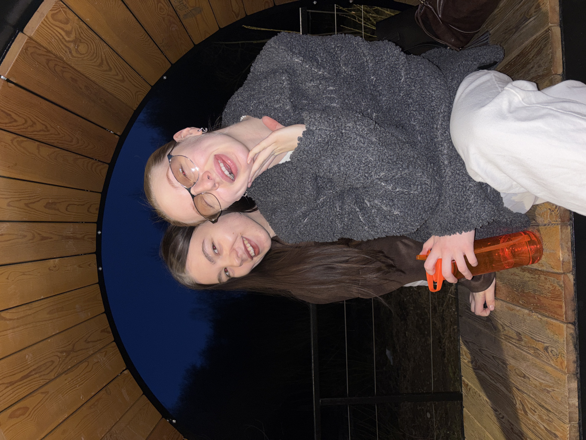 Two young women are posing for a picture, possibly a selfie, in what appears to be a wooden circular structure. The woman on top has fair skin, light hair, and is wearing glasses, a gray fuzzy jacket, and white pants. The woman below has darker skin and long dark hair, and is holding an orange water bottle in her hand. The structure is made of curved wooden planks, and the background appears to be a dark, outdoor scene, possibly near a body of water or forest. The location may be in Perm, Russia.