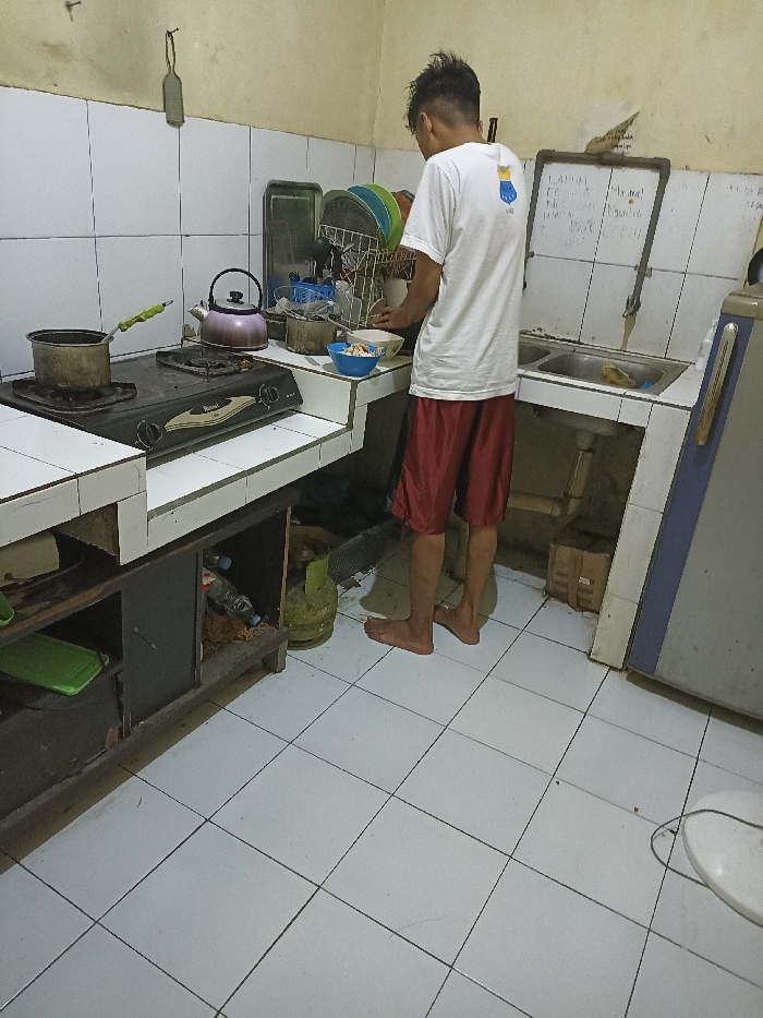 The image depicts a kitchen interior, located in Jatinangor, Indonesia, at coordinates -6.934 latitude, 107.779 longitude. A young man, wearing a white T-shirt and red shorts, is standing near a gas stove and kitchen sink. The man is facing the sink and is looking to his left. He is holding a white bowl in his left hand and has his right hand on the edge of the countertop. The stove is on the left, with two burners and a black metal grate. A red kettle, a green tea kettle, and other kitchen utensils are on the counter next to the stove. The sink is to the right of the man and is made of stainless steel. It has two basins, one of which is partially filled with water. The kitchen has white tiled walls and a white tiled floor. There is a black cabinet with a wooden shelf beneath the countertop. Several items are on the shelf, including a bottle of liquid and a green gas cylinder. A large white refrigerator is in the background. The kitchen appears to be in a state of disarray, with some items out of place and some dust on the floor.