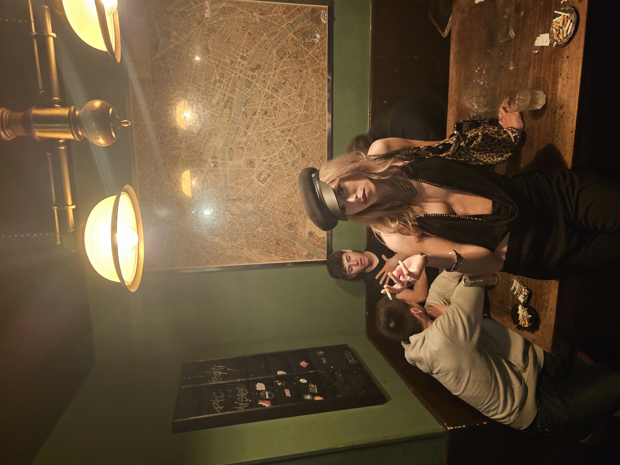 The image shows a dimly lit bar in what appears to be Paris, France, evidenced by a large map of the city on the wall. Three people are visible: a blonde woman wearing a black hat, a black top and an animal print bag and two men sitting at a wooden table. The woman is looking towards the camera, while one of the men is smoking and the other is turned away. The table is cluttered with cigarette butts, a glass, and a pack of cigarettes. A light fixture with three globes hangs above, and a menu board with writing and stickers is mounted on the green wall.