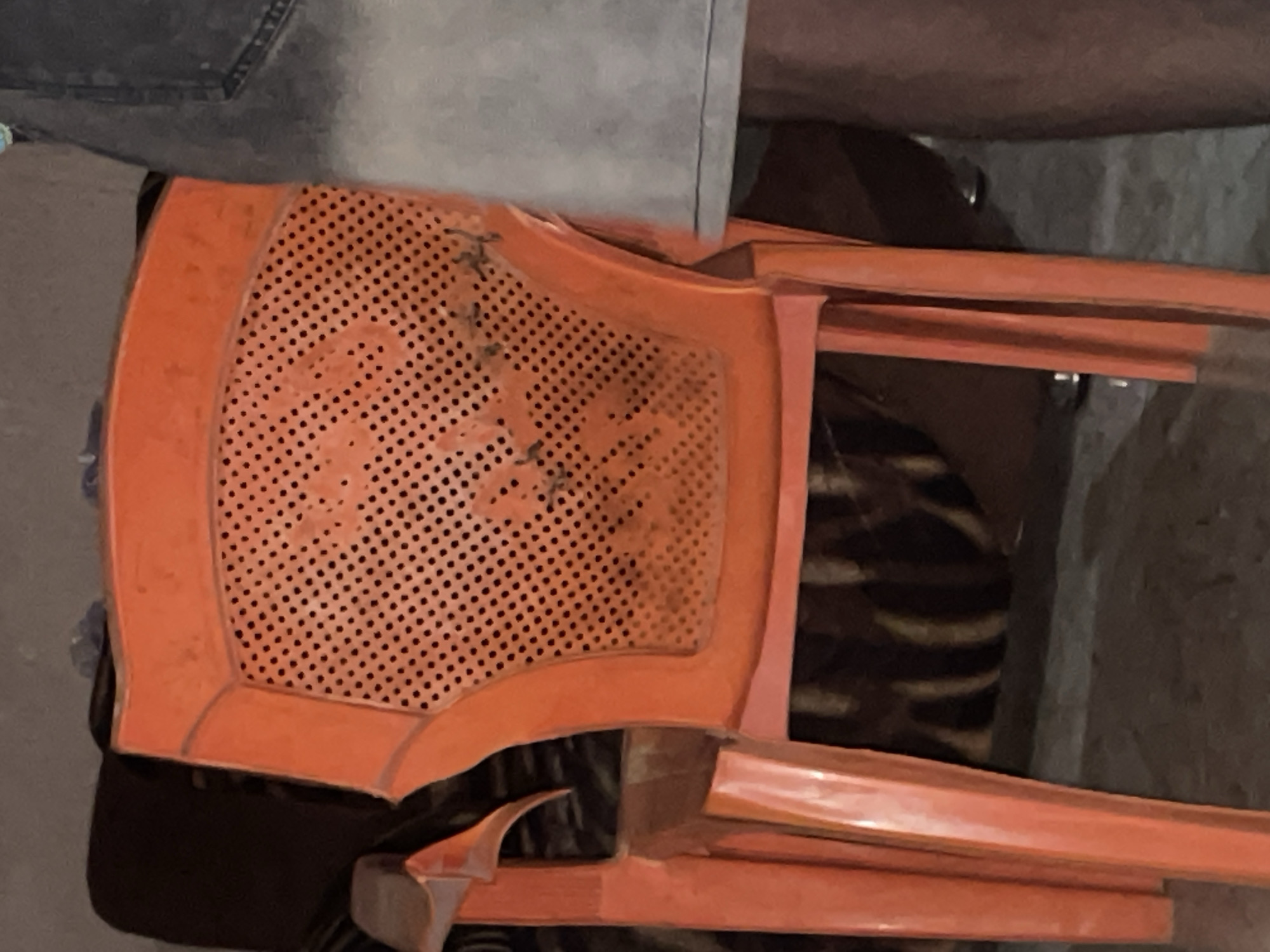 The image shows an orange plastic chair with a perforated backrest. There are dark marks or stains on the backrest. To the left of the chair, part of a black couch or armchair is visible. To the right of the chair, a person's legs wearing denim jeans are visible. The scene appears to be indoors, possibly in a home or a common area. The lighting is dim, suggesting it might be evening or an interior space with limited natural light. The floor is concrete or a similar hard surface. The overall impression is of a simple, functional setting. Without further information, it is impossible to determine the specific location within Lokoja, Nigeria.