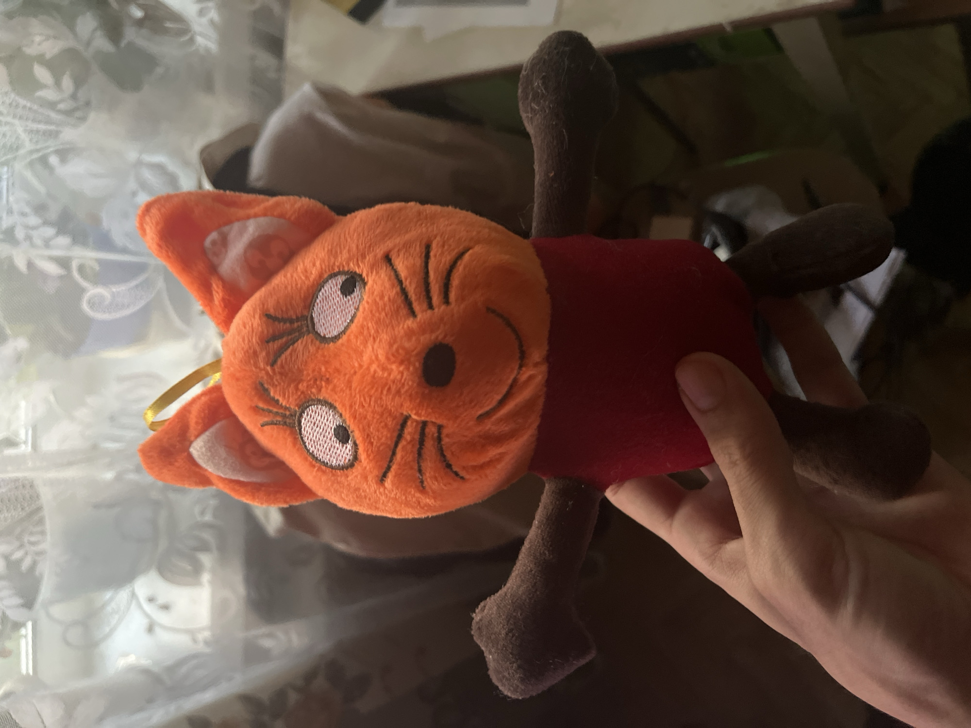A person's hand is holding an orange plush toy fox with dark brown arms and legs. The fox has a red body and a cartoonish face with wide white eyes with black pupils, thin black eyebrows, a black nose, and a smiling mouth. It has two pointy ears with white inner ear details. A yellow ribbon is attached to the top of its head. The background is blurred but appears to be indoors with a window covered by white lace curtains. There are some indistinct objects visible behind the toy, including what might be a white teacup and saucer on a shelf. The lighting suggests an indoor environment, possibly a domestic setting. The image does not contain any identifiable landmarks or features that would indicate a specific location in Kazan, Russia.