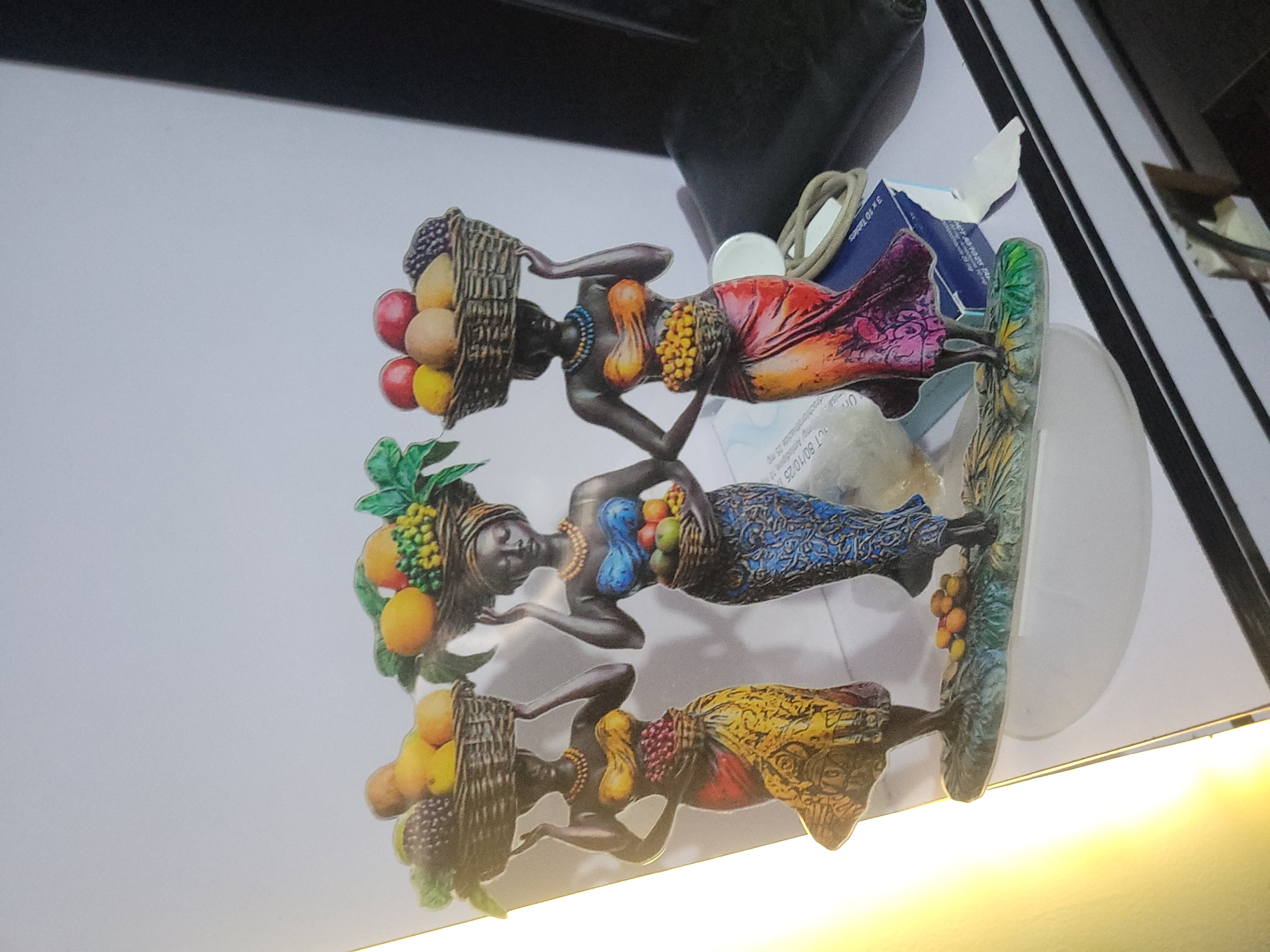 The image shows a decorative figurine of three dark-skinned women, each carrying a basket of fruit on their head and another smaller basket of fruit at their waist. The women are adorned with colorful garments and beaded necklaces. The figurine is placed on a white surface with a white background. To the right of the figurine, a dark object, possibly a piece of furniture or electronic device, is partially visible. The overall lighting suggests an indoor setting. The provided information states this scene is located in the city of Jos, Nigeria.