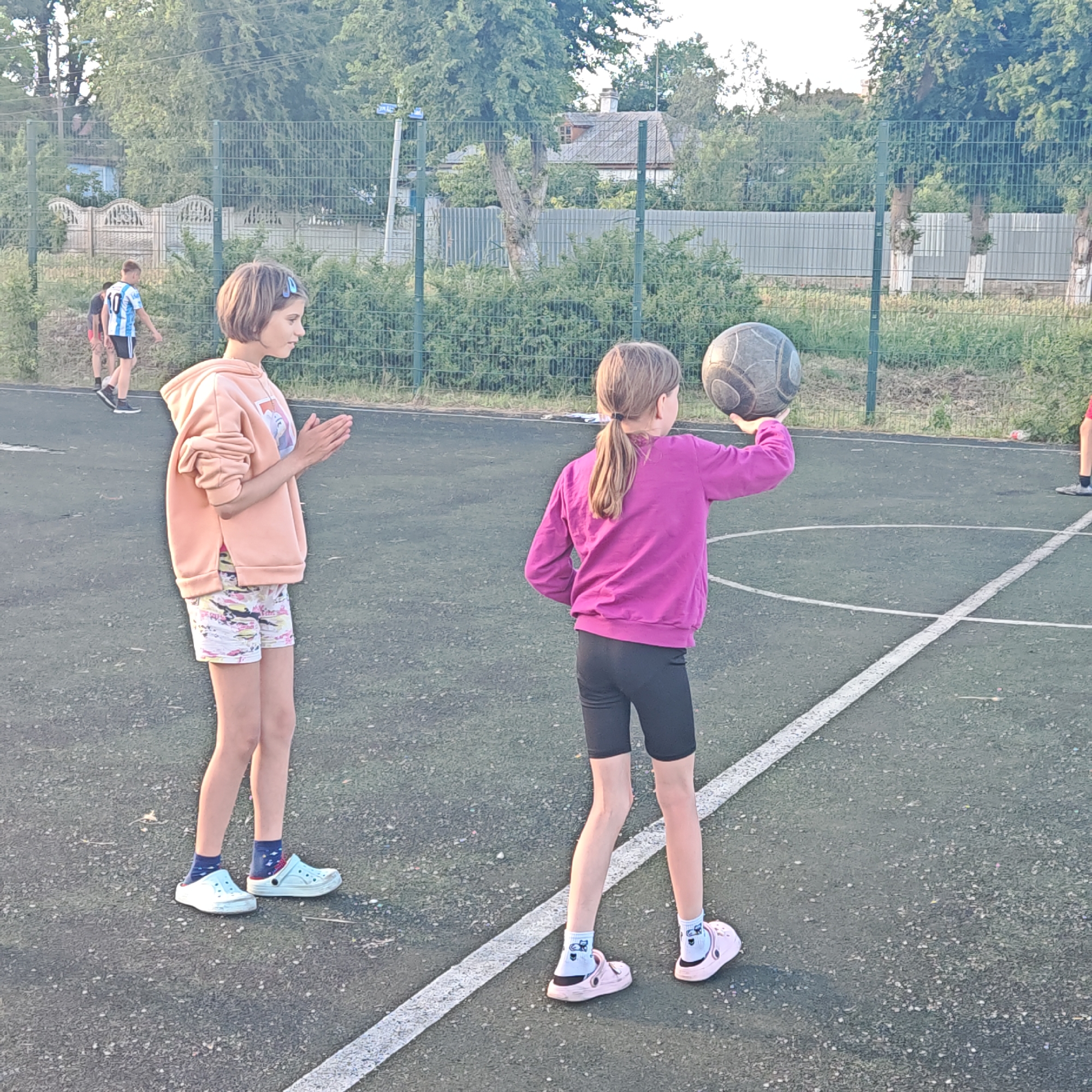 The photograph depicts a sunny outdoor scene in Мысовское сельское поселение, Ukraine. Two young girls are on an asphalt sports court. One girl, wearing a peach-colored hooded sweatshirt and patterned shorts, stands with her hands clasped, seemingly clapping. The other girl, facing away from the viewer, wears a purple sweatshirt and black shorts; she holds a worn, dark-colored ball in her raised hand, preparing to throw it.
In the background, a chain-link fence runs along the perimeter of the court, with trees and greenery visible beyond. A glimpse of a house roof and the top of a white fence can be seen through the trees. A boy in a sports jersey is walking in the distance on the left side of the image, and a portion of another person is visible on the right edge of the image. White lines mark the boundaries of the court.