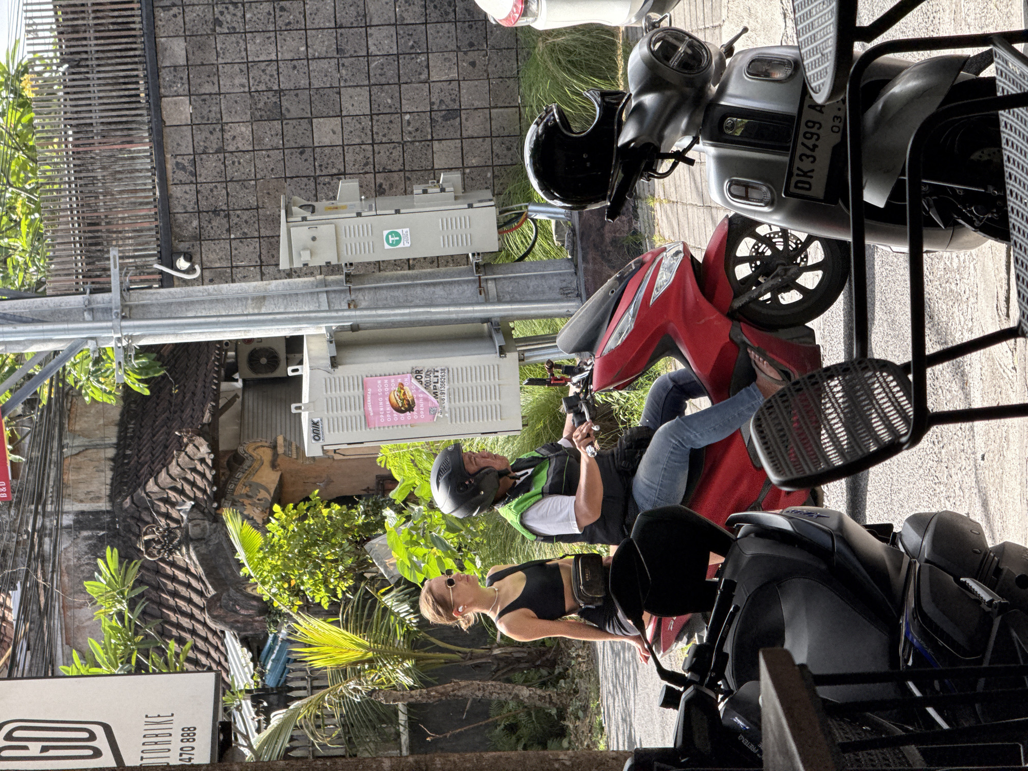 A street-level, rotated, medium shot shows a partially obstructed view of a red scooter and a silver scooter parked on the side of a street. A man and woman are on the red scooter. The man is driving. He wears a black helmet, a vest with white and green panels, and blue jeans. The woman is in front, wearing a black halter top, black shorts, sunglasses, and a small black bag around her waist. The scooter and the people on it are facing left. Another scooter, gray and black, is in front of them and to the right. At the top, the license plate of the silver scooter is visible and reads "DK 3499 A." Behind the scooters, a pole supports two electrical boxes and a street sign on top. The lower box has a sticker partially visible that reads, "BURGERHEAD OPENING SOON OPENING SOON." A sign with the word "OTORBIKE" is at the bottom left. The street is concrete. Green foliage is behind the people and vehicles. The location appears to be North Kuta, Indonesia.