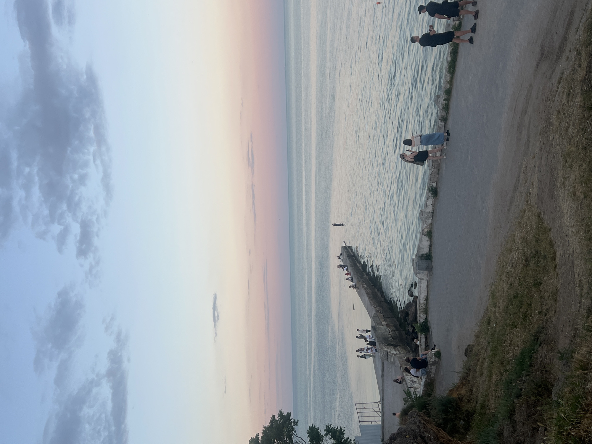 The image shows a pier extending into a calm body of water, likely a sea, under a twilight sky. The sky displays a gradient of soft blues and pinks. Several people are scattered along the pier, some sitting and others standing, appearing to enjoy the view or fish. On the shore, a paved pathway runs parallel to the water, where a few more individuals are visible, some walking and others conversing. The foreground features a grassy embankment sloping downwards towards the path. The overall scene depicts a serene, late evening gathering by the water.