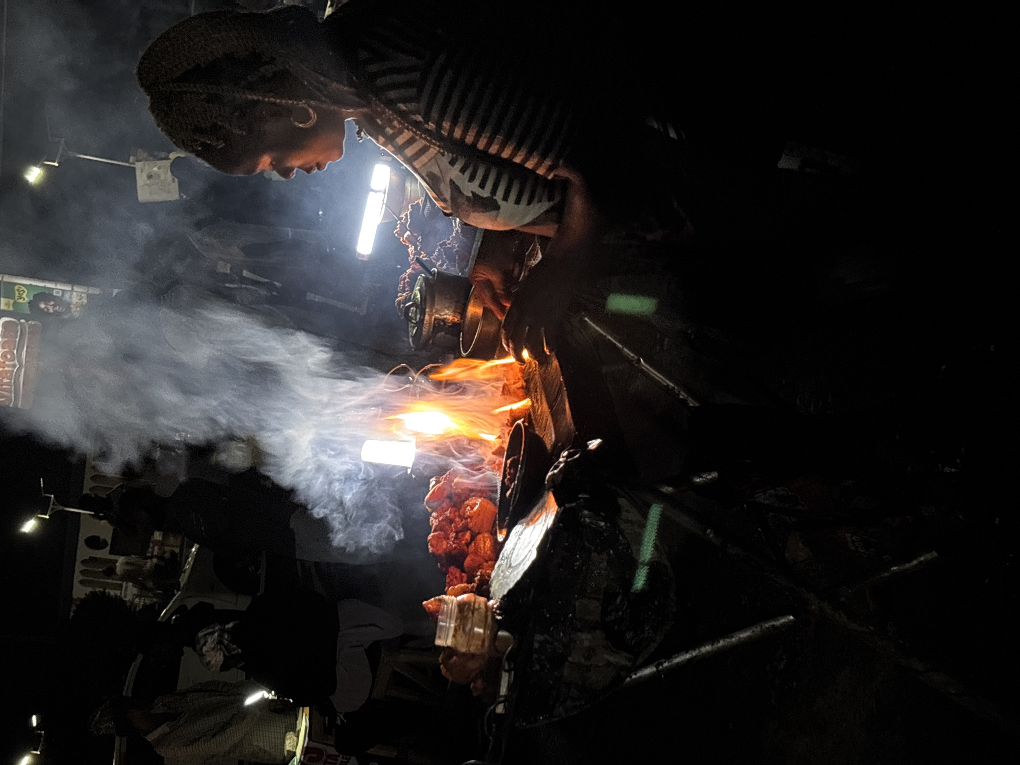 A low-angle shot depicts a woman standing in front of a meat spit, likely a shawarma or suya stand, emitting smoke and flames, suggesting food is being prepared. The woman, wearing a striped top and a hat, is illuminated by bright lights. The location is the city of Jos, Nigeria. The overall environment appears dimly lit, with other objects and figures visible in the background, though obscured.