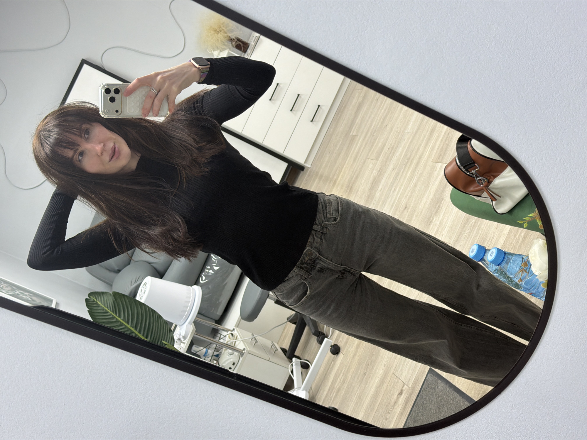 A woman with long, dark hair and bangs is captured in a mirror selfie, taken indoors in Perm, Russia. She is wearing a black ribbed long-sleeved top and dark grey, wide-leg jeans. Her right hand holds a light-colored smartphone with a clear case, positioned to take the picture, while her left hand is raised, gently touching her hair. She has a ring on her right ring finger and a smartwatch with a colorful strap on her left wrist.
The setting, reflected in the dark-framed mirror, features light wooden flooring and white walls. In the background, a white cabinet with three black vertical handles is visible, topped with a fluffy decorative item. Parts of a black-framed picture or mirror are also reflected. To her left, a light grey armchair or sofa, a white floor fan, and a large green artificial plant leaf are seen. To her right, a brown and white handbag rests on a green surface, alongside two blue plastic water bottles. The bottles clearly display the brand name "AQUA" and the Russian text "АРТЕЗИАНСКАЯ ВОДА" (Artesian Water). A white rose or flower is also present near the bottles. The reflection also shows parts of an office chair and a small white rolling cart, suggesting a modern workspace or home office environment. The woman maintains a soft, neutral expression looking directly into the mirror. The space appears brightly lit, indicating either daytime or good indoor lighting.