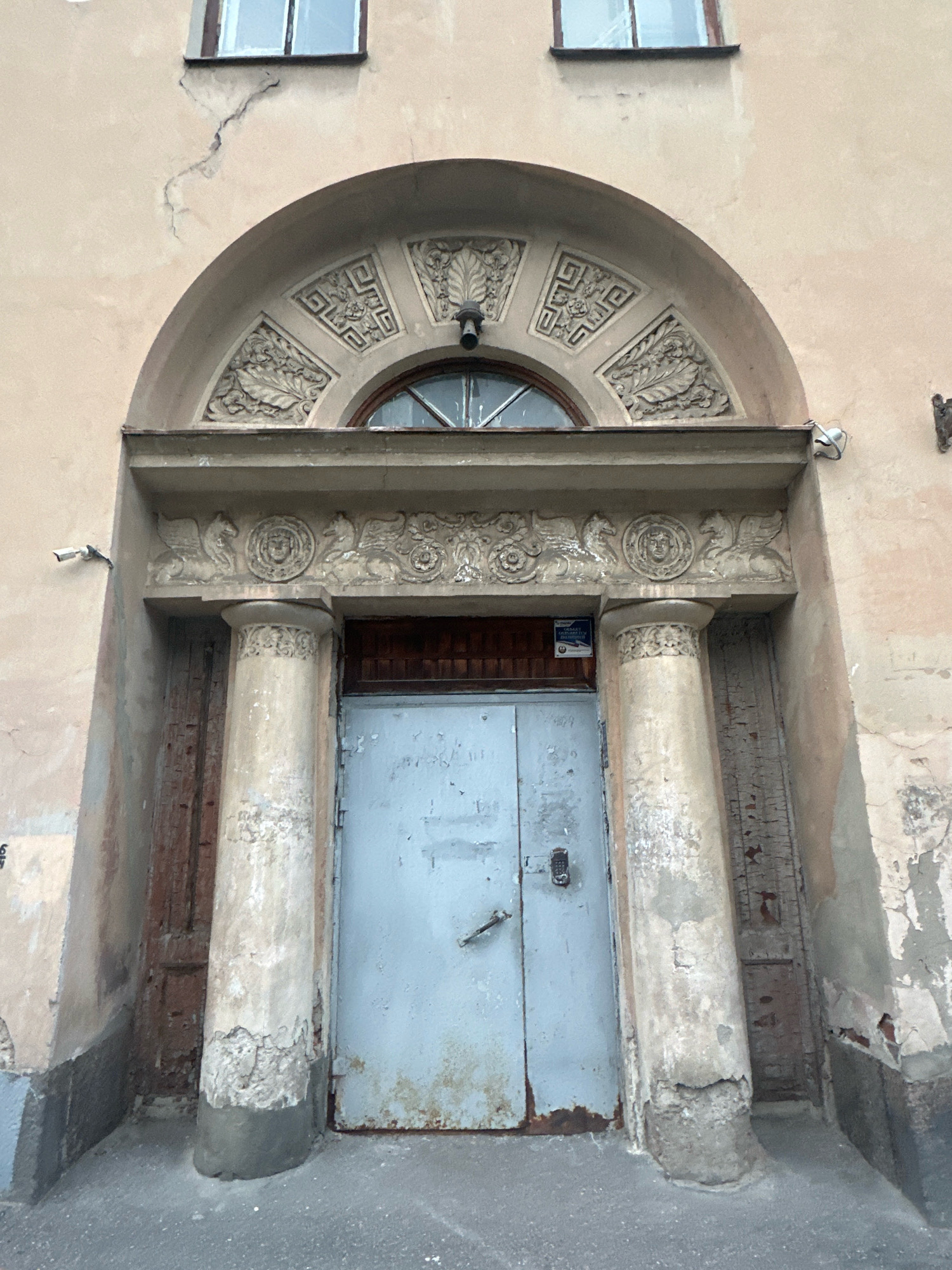 The image shows a close-up of a weathered, pale pink building facade with architectural details, likely in St. Petersburg, Russia. Dominating the center is a large, pale blue double door with a metallic handle and a digital lock. The door is flanked by two fluted columns with distressed, textured surfaces, resting on rough, grey concrete bases. Above the columns is an ornate stone entablature featuring relief carvings of griffins on either side of a circular medallion with a face. This entablature supports a decorative archway, also adorned with relief carvings of leaves and geometric patterns, framing a semicircular window with a visible pane. Two white security cameras are mounted on the facade, one on the left side above the columns and another on the right above the entablature. Cracks and peeling paint are evident on the building's walls, especially around the window and columns, suggesting age and exposure to the elements. The ground in front of the doorway is paved with asphalt. A small sign, possibly an information or warning sign, is affixed to the wall to the left of the door.