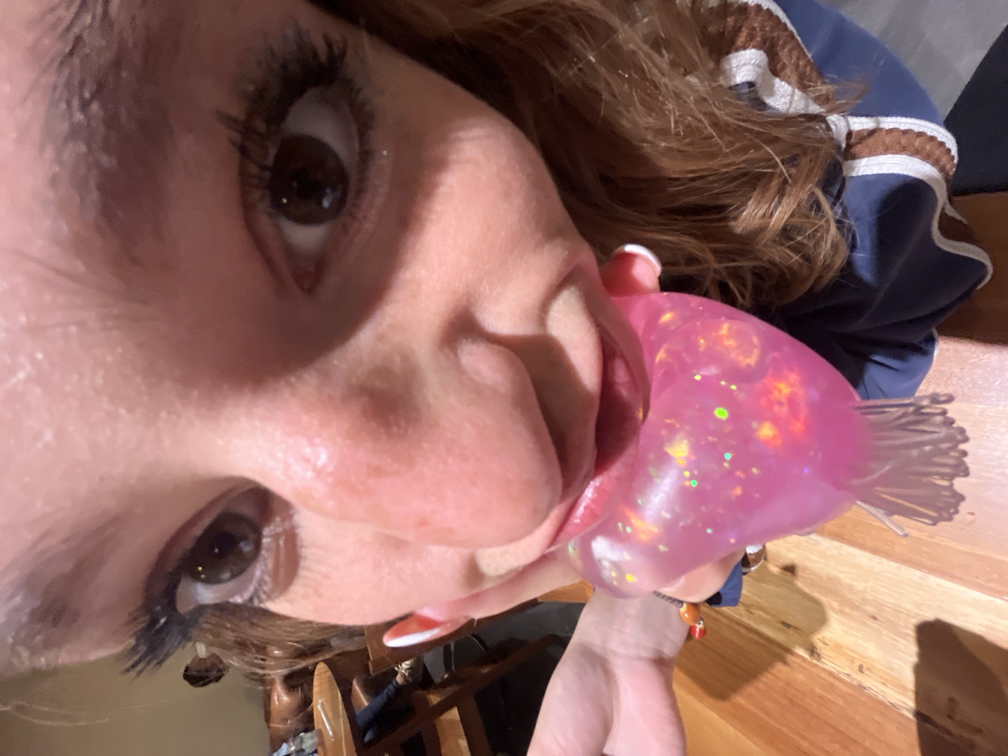 A close-up, tilted, and slightly distorted image shows a person with brown eyes, fair skin, and long eyelashes holding a pink, glittery light-up toy near their mouth. The person has a visible white nail polish. The toy is glowing and features a bundle of thin, clear, protruding fibers. The background appears to be a wooden surface, possibly a table or floor. Part of a blue garment with white and brown striped trim is visible on the right side of the frame. The location is unknown.