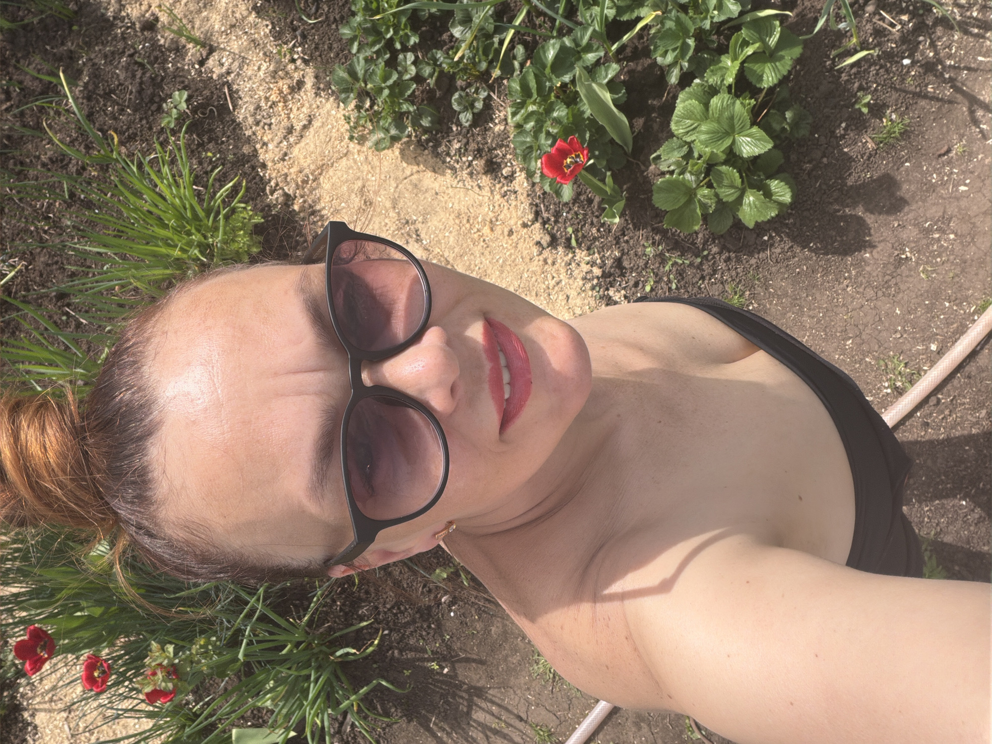 The image is a selfie of a woman with her hair in a bun, wearing sunglasses and lipstick, in an outdoor setting, possibly a garden. The background includes soil, grass, a red tulip, and plants that appear to be strawberry plants. A garden hose is visible in the background. It is impossible to ascertain that this photo was taken in Chaikovsky, Russia.