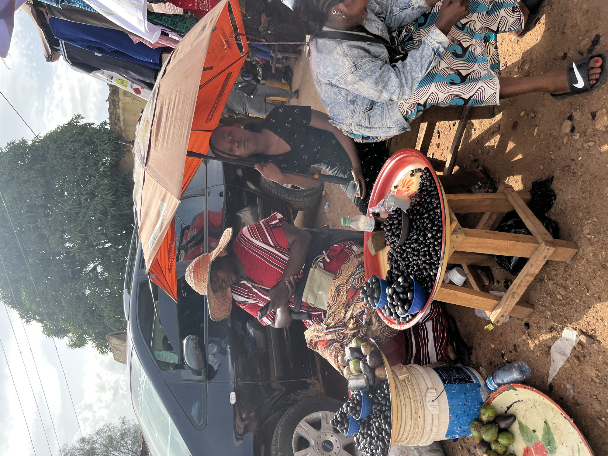 This media file captures a street market scene in Rafin Par, Nigeria. The primary subjects are three women, two of whom appear to be vendors and one a customer. One vendor, wearing a black shirt with a subtle pattern, is seated under an orange umbrella, looking towards the camera. Beside her, another woman, wearing a light blue denim jacket and a patterned blue and white dress, is also seated. The third woman, dressed in a red and white striped top and a straw hat, is actively engaged, possibly arranging produce.
The scene is set outdoors on a dusty ground, with a dark-colored vehicle parked behind the women, serving as a backdrop and likely a mode of transport. Scattered around are various objects related to the market activity: buckets and bowls filled with dark berries, possibly olives or a similar fruit, and a separate bowl containing green fruits, which appear to be avocados. A plastic water bottle and other assorted market debris are also visible.
The activity appears to be a typical street market transaction, with the vendors selling their goods. The time of day seems to be daytime, indicated by the shadows cast on the ground and the bright lighting. The weather appears to be clear and sunny. The overall atmosphere is that of a lively, informal marketplace. There are no discernible emotions beyond casual engagement.
A partially visible orange flag or banner extends from the umbrella, featuring some unreadable text. The car's wheel is visible, with "FORD" text on the hubcap.