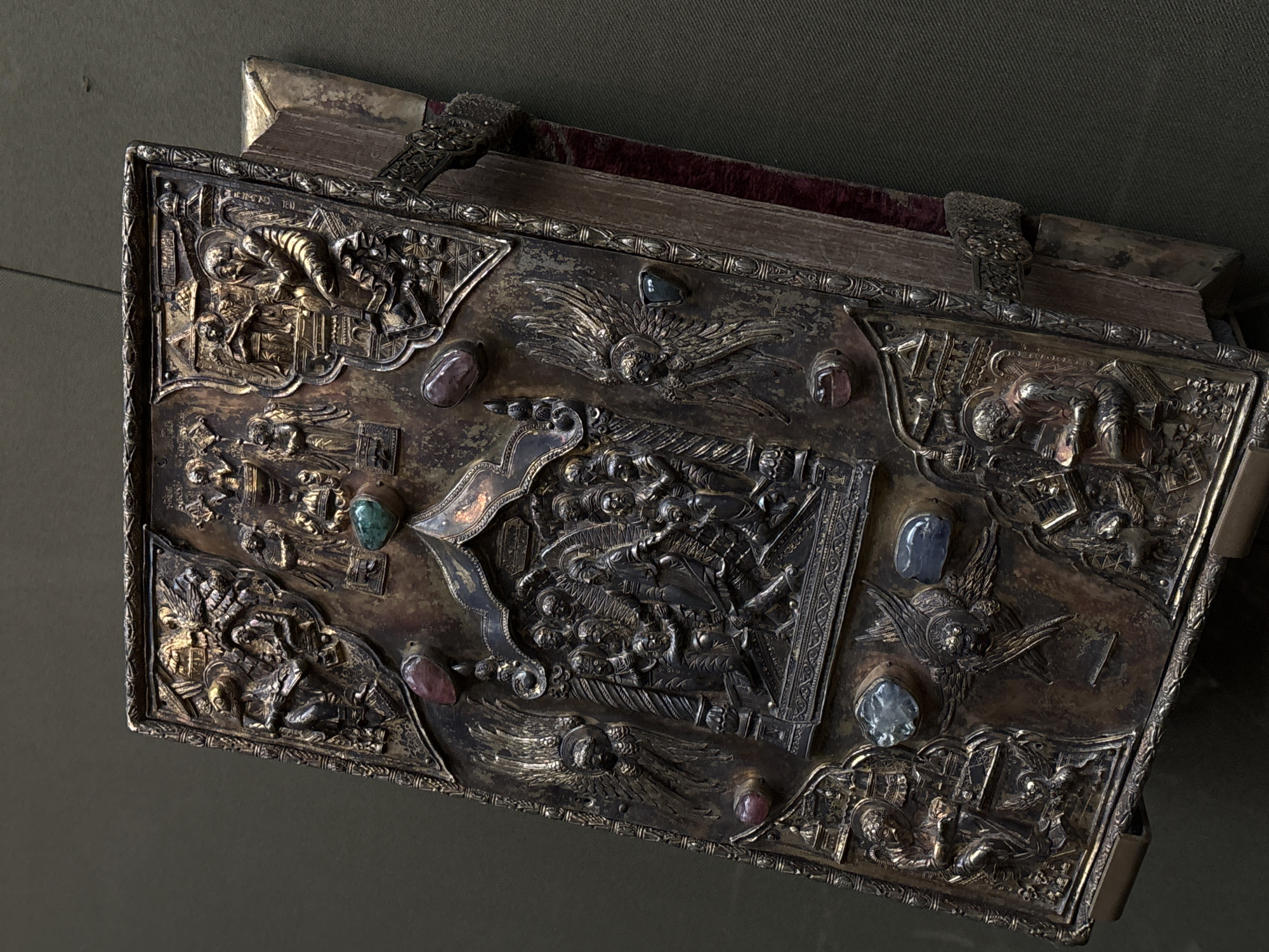 The image shows a highly ornate, antique book with a metal cover. The cover is decorated with relief sculptures of figures and scenes, along with several gemstones. The book appears to be resting on a dark, possibly velvet-covered surface, adding to the impression of historical significance and value. The style suggests religious art or a historical artifact.
Without further context or labels, it is impossible to confirm its specific location in Moscow, Russia or provide details of the subjects depicted on the cover.