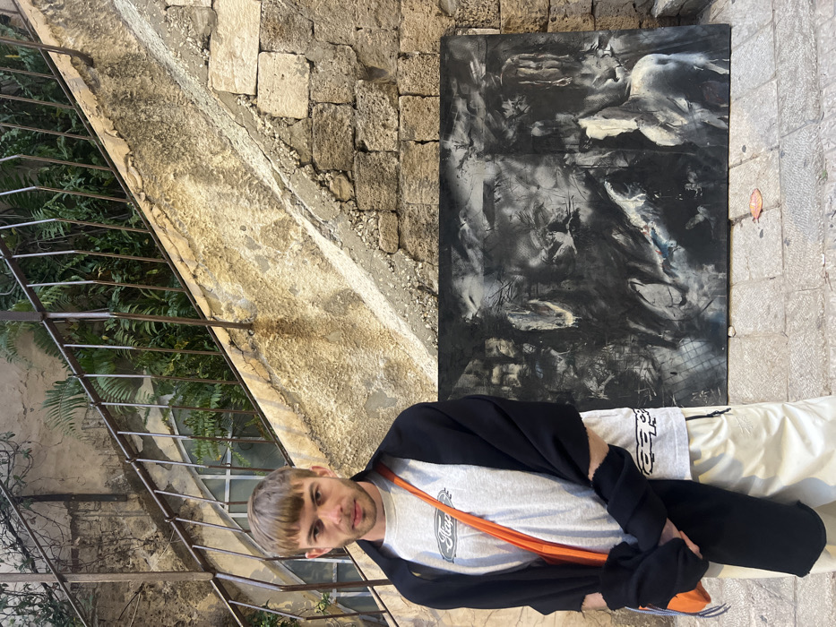 The image captures a full-length portrait of a man standing outdoors next to a large abstract painting. The photo appears to have been taken in a vertical orientation but is currently rotated 90 degrees clockwise, placing the man on his back and the background sideways.
In the foreground, a light-skinned man with short, light brown hair and bangs is looking directly at the camera with a neutral expression. He is wearing a light grey t-shirt with a dark logo, which appears to be the word "Staple" with a small bird or pigeon design above it. An orange strap, possibly from a bag, crosses his chest. A black garment, possibly a shawl or jacket, is draped over his shoulders and arms, with one sleeve partially covering his left hand. He is also wearing light-colored, possibly white or cream, trousers.
To the man's right (which would be behind him if the image were properly rotated), a large, dark-toned abstract painting is propped against an old stone wall. The artwork is predominantly black and white with shades of grey, featuring broad, expressive brushstrokes and smudges. Lighter, almost ghost-like shapes emerge from the dark background; one form in the upper right quadrant resembles a crouching figure or perhaps part of an animal, while other white and grey elements create a sense of dynamic movement and texture across the canvas.
The background consists of several elements. To the man's left (foreground left), there is a metal railing, possibly part of a staircase or balcony, with lush green foliage, likely ferns, visible through and behind it. Behind the man and the painting, an old wall constructed from large, irregularly shaped, light-colored stone blocks is visible. The ground is paved with similar stone or concrete. The overall setting suggests an ancient or rustic urban environment, possibly in a historical district or a Mediterranean locale, given the stone architecture and visible greenery. The lighting indicates it is daytime, casting soft shadows.