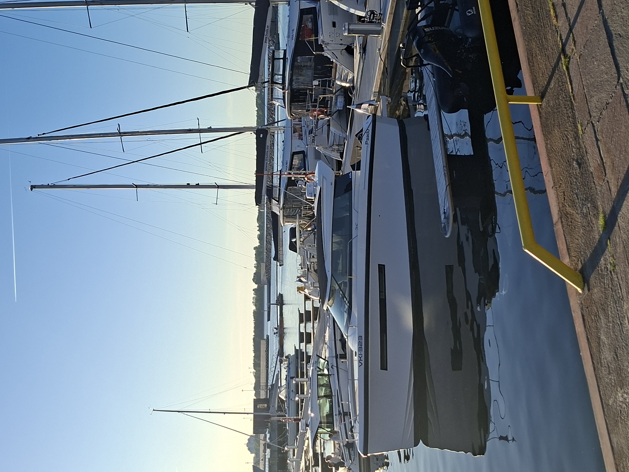This detailed image depicts a marina in Tallinn, Estonia, under clear daytime conditions with a low sun angle suggesting either early morning or late afternoon light. The sky is a gradient of light blue at the horizon, transitioning to a deeper blue overhead, with a faint white contrail horizontally bisecting the upper third of the frame.
In the immediate foreground, a textured, reddish-brown paved dock made of rectangular stone blocks extends from the lower right. Small patches of green vegetation are visible in the cracks of the paving. A bright yellow metal railing with a horizontal top bar and a vertical support post occupies the lower right corner, casting a shadow towards the left.
The calm, dark blue water of the marina fills the majority of the lower half of the image, reflecting the boats and sky with minimal ripples. Numerous boats are docked in parallel, extending from the mid-ground towards the distant background.
The most prominent subject is a modern, white powerboat positioned in the left mid-ground, featuring a dark hull stripe below the window line. The registration "VKJ-323" is displayed in black on its bow, and the name "Paadu" is visible on its port side. A small Finnish flag is attached to the stern of this boat.
Behind this powerboat, other similar white powerboats are docked. One visible registration on a boat further back is "VKA-951". Another powerboat in the right mid-ground displays "VKH-156" and "400 S2" on its stern. Several sailboats are also present, distinguished by their tall, slender masts and rigging. The boom covers of multiple sailboats bear the web address "www.5sails.ee" in white text, indicating an Estonian yacht charter company. Two individuals are visible seated in the cockpit area of a powerboat docked directly behind the foreground vessel. In the water below the yellow railing, an inflatable dinghy with a "Suzuki" outboard motor is partially visible and reflected.
In the distant background, the shoreline of Tallinn is visible, featuring various industrial or port structures, possibly including cranes or warehouses, and a line of trees. The lighting creates a slight atmospheric haze towards the horizon.