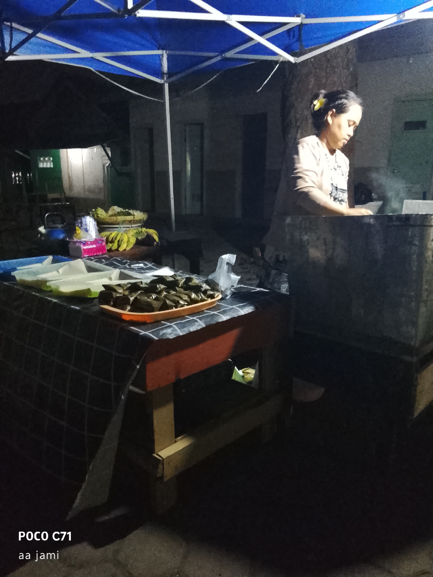 The image depicts a street food stall at night in Tasikmalaya, Indonesia. A woman with her hair tied up, adorned with a yellow flower, is standing behind a table laden with food items, including what appears to be traditional Indonesian snacks wrapped in banana leaves. She is wearing a light-colored long-sleeved shirt. In front of her, there are plastic containers holding more food, and a basket of bananas. The stall is covered by a blue and white tent, and the surrounding environment is dimly lit, suggesting it is a nighttime scene in an urban or semi-urban setting. The watermarks "POCO C71" and "aa jami" are visible in the bottom left corner.