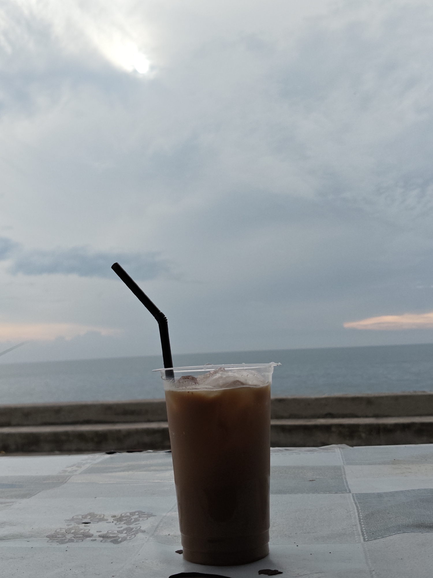 A clear plastic cup filled with a light brown beverage and ice cubes, with a black straw inserted, is positioned on a table with a checkered pattern. The cup is in the foreground. In the background, a calm body of water, presumably the sea, stretches towards the horizon. Above the water, a cloudy sky is visible, with the sun partially obscured by clouds, casting a bright spot in the upper left portion of the frame. The overall lighting suggests it is either early morning or late afternoon. The scene is likely outdoors, possibly at a cafe or dining area near the coast in Lhokseumawe, Indonesia, given the presence of the sea and the style of the outdoor table. No people are visible in the image.