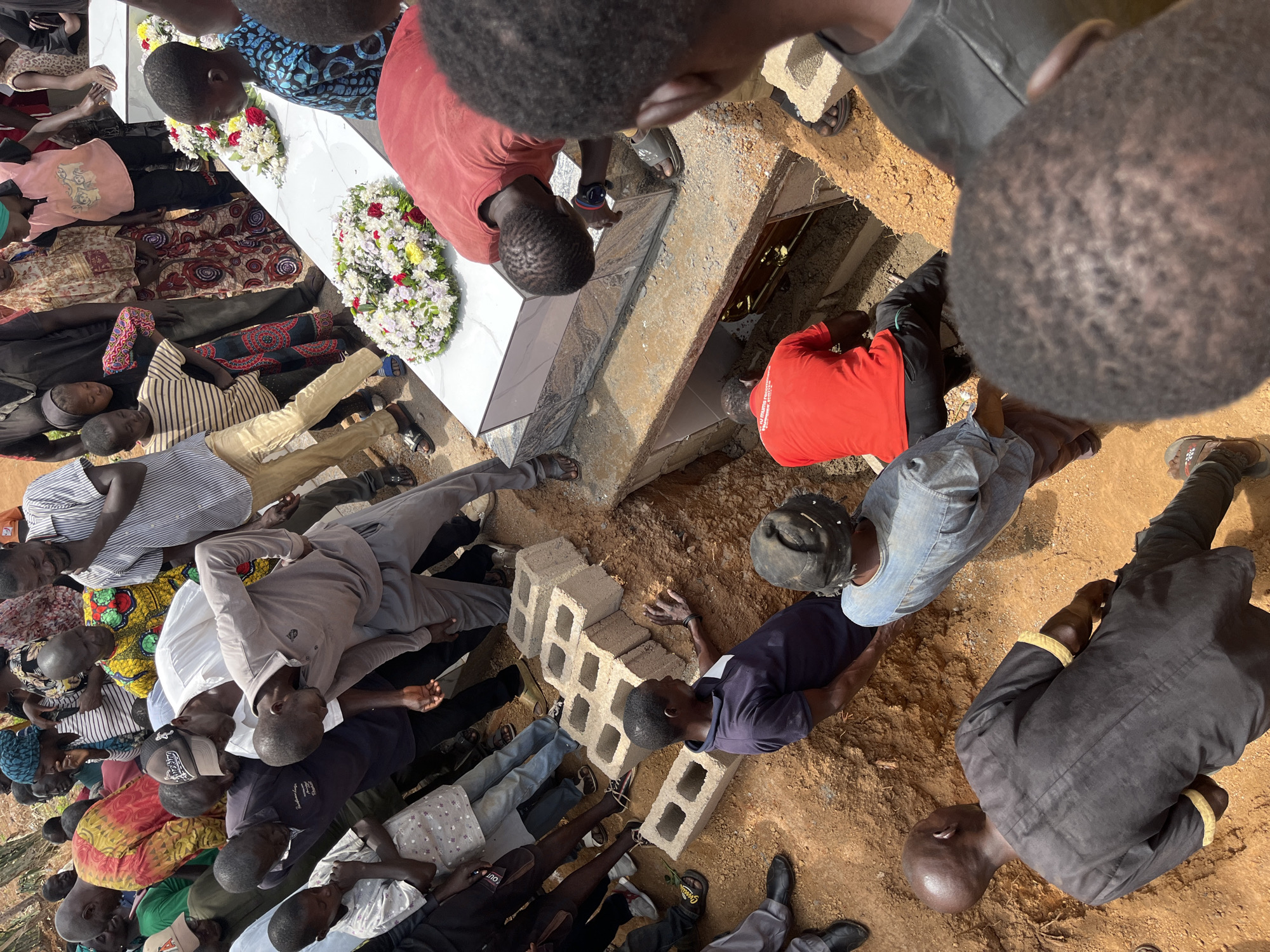 The image shows a funeral taking place in Dutsen Kato, Nigeria. A large group of people surrounds a grave site where a white coffin lies on top of the opened grave. Two floral wreaths are placed on the coffin. Two men in red shirts are working within the grave to prepare it, while others stand around the perimeter, some on cinder blocks. The ground around the grave is sandy.