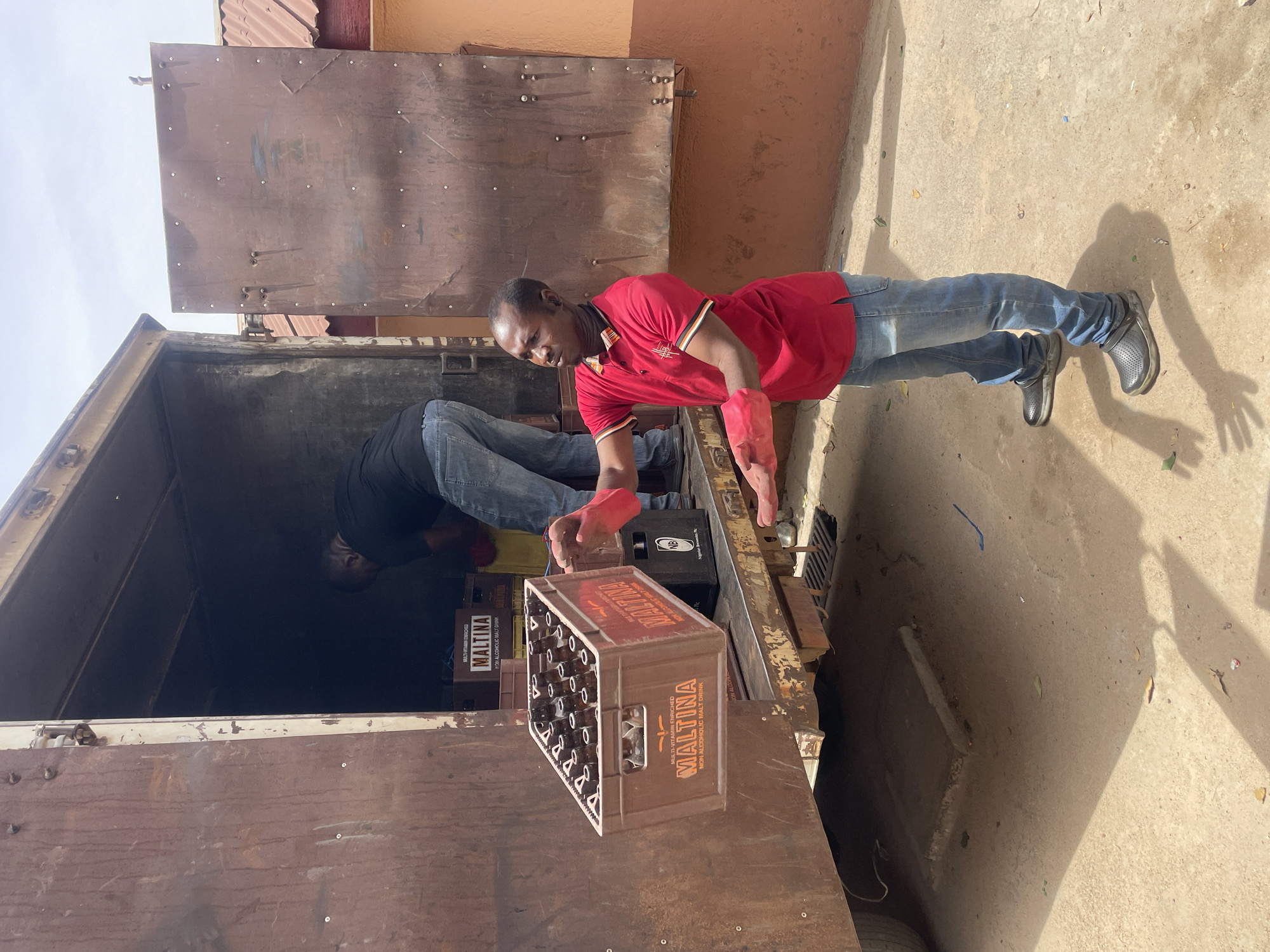 This outdoor photograph, taken in Jos, Nigeria, captures two men engaged in the loading or unloading of beverage crates from a delivery vehicle. The scene is illuminated by bright daylight, casting strong shadows on the concrete or dirt ground, suggesting a mid-day sun.
The most prominent subject is a man in a red polo shirt, blue jeans, and dark clogs, wearing red gloves. He is positioned at the back of an open-sided utility or delivery vehicle, actively handling a brown plastic crate filled with bottles. Another man, wearing a dark shirt and blue jeans, is partially visible inside the dark cargo area of the vehicle, also involved in the task.
The crates are distinctly branded "MALTINA," with additional text "MULTI-VITAMIN ENRICHED" and "NON ALCOHOLIC MALT DRINK." One black crate visible inside the vehicle also shows "NB" and appears to include "Nigerian Breweries Plc." The vehicle itself appears sturdy and well-used, characteristic of a working delivery truck. In the background, there's a building featuring an ochre-colored wall and corrugated metal siding. The men appear focused on their work, and the overall atmosphere is one of routine commercial activity.
