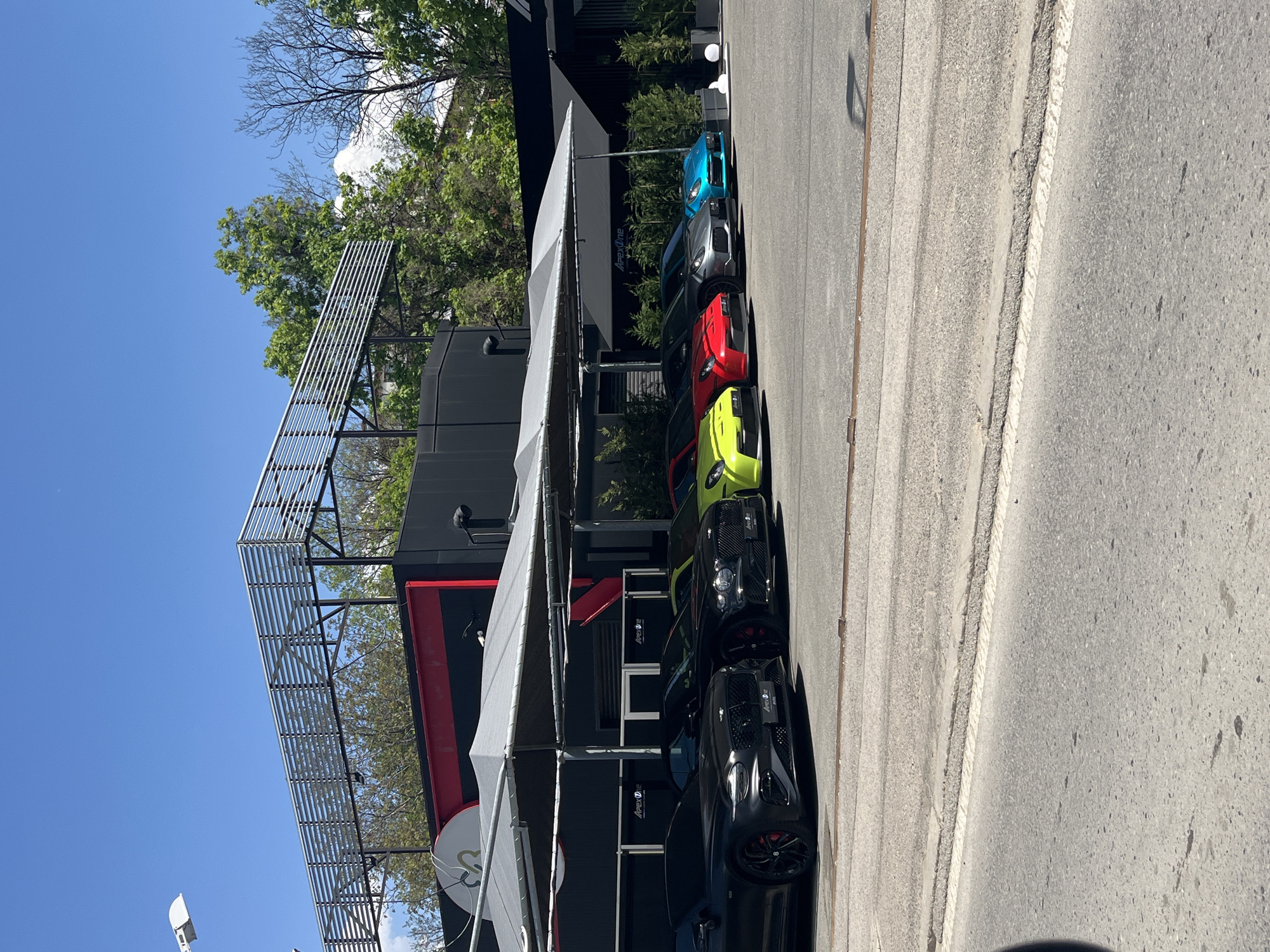 The image depicts a row of luxury cars parked adjacent to a building with a metal framework structure overhead. The cars are various colors, including black, green, red, silver, and blue. The building has black and red accents. A white awning extends over the parking area. The location is in Sofia, Bulgaria as evidenced by the signage "ApexOne" visible on the building. The street is paved with a light grey concrete. The sky is clear blue with trees visible in the background. No people are visible in the frame.