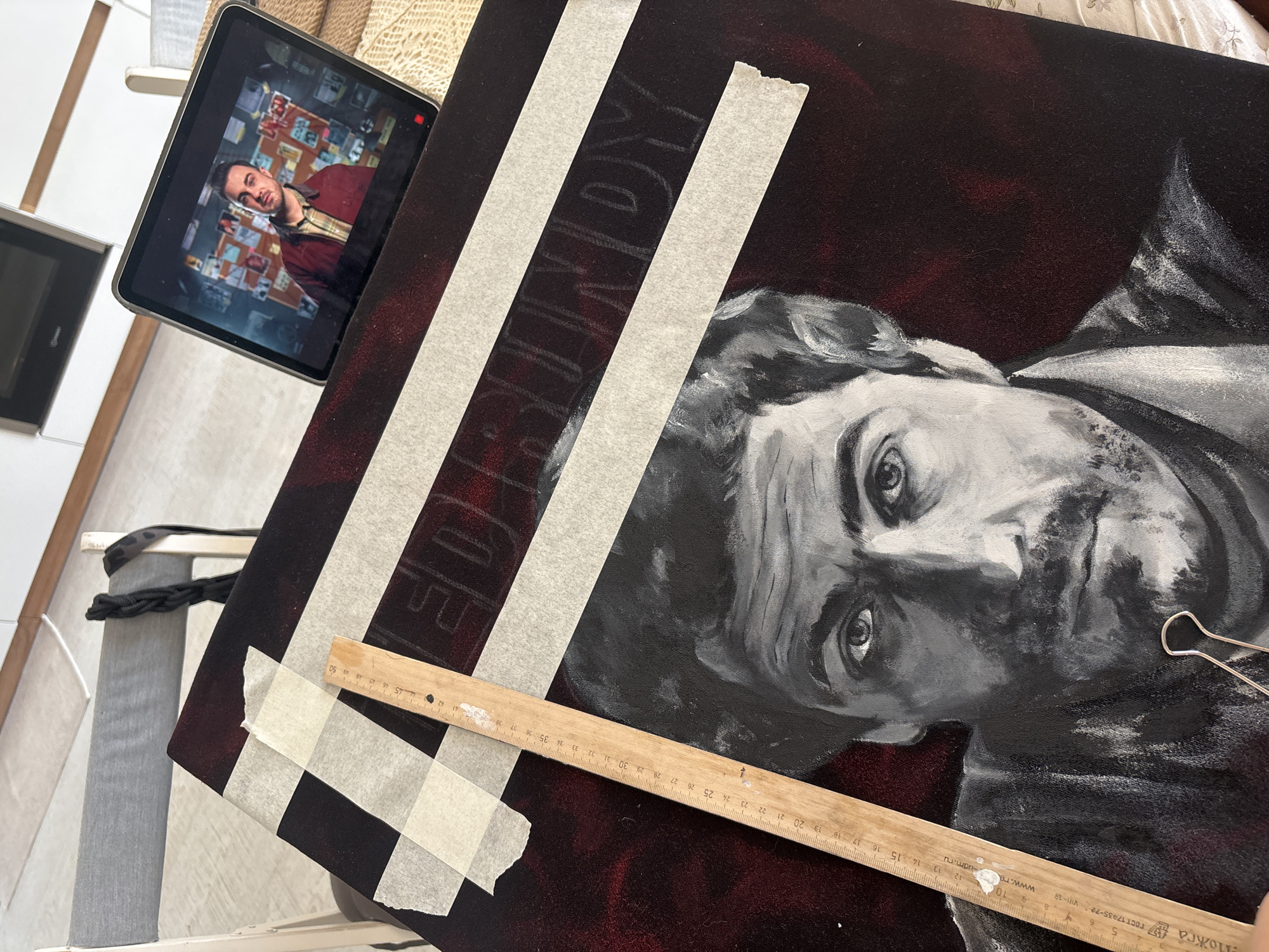 The image shows an in-progress painting of a man in greyscale, likely based on the image displayed on the tablet next to it. The background of the painting is a deep burgundy color. White masking tape is used to form stripes on the canvas, with lettering underneath the tape. A wooden ruler lies across the painting, with a small amount of white paint on it. A paperclip is also near the painting. The work area appears to be indoors. It is not possible to determine a location in Krasnoyarsk, Russia from this image.