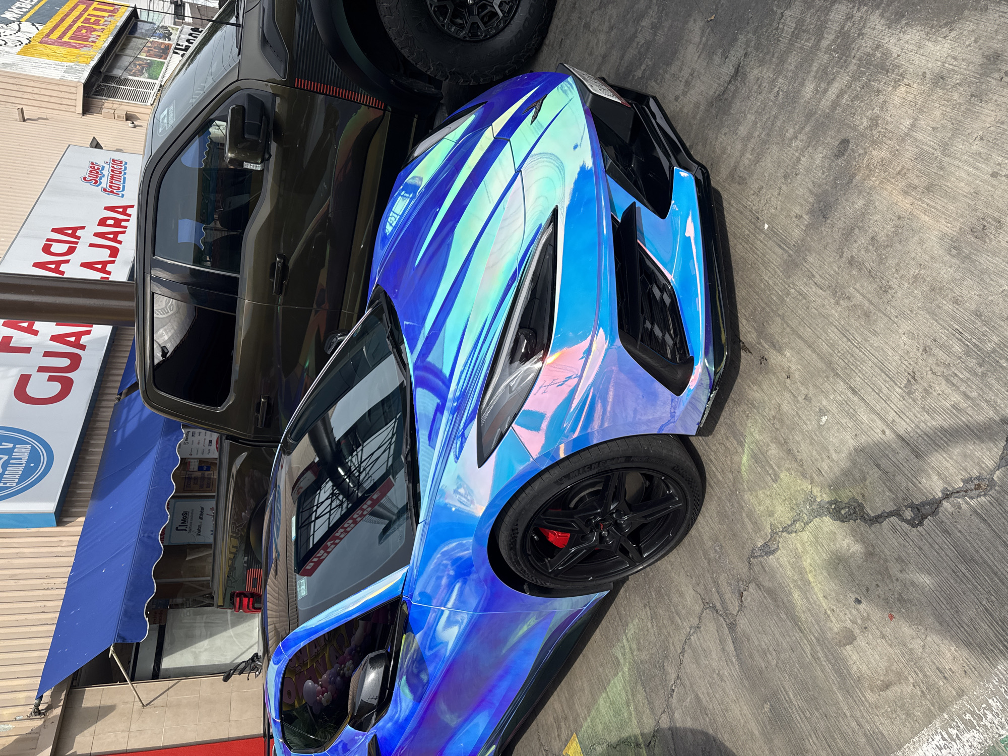 The image depicts a shiny, iridescent blue sports car parked on a gray concrete surface. The car is positioned next to a brown pickup truck. In the background, there's a building with an awning and a sign for "Super Farmacia Guadalajara", indicating the location is likely in Puebla, Mexico. The photo appears to be taken during daylight hours. The vehicles seem to be stationary.