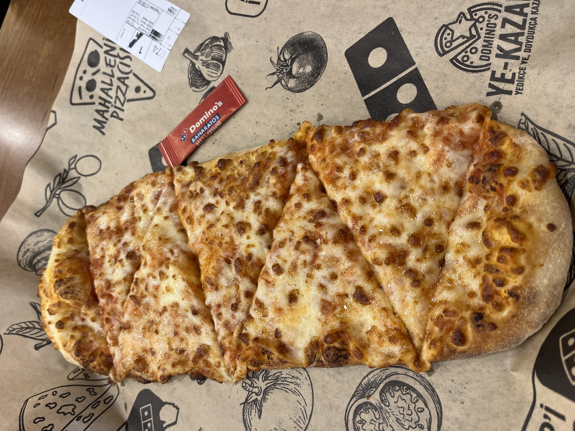 The image shows a sliced Domino's pizza on branded wrapping paper. The pizza is topped with cheese and sauce. A small packet of Domino's "Baharatos Pizza Cesnisi" (seasoning) is placed near the pizza. The wrapping paper has images of tomatoes and garlic printed on it, as well as the Domino's logo and Turkish text "Yedikce Ye, Doydukca Kaza" and "Mahallen Pizzacisi." A receipt is placed at the corner of the pizza box. The receipt includes the text "Toplam TL120.00" and lists prices and tax amounts for items that total "TL109.09, TL10.91, and TL3.30". The pizza appears to be in Alanya, Turkey.