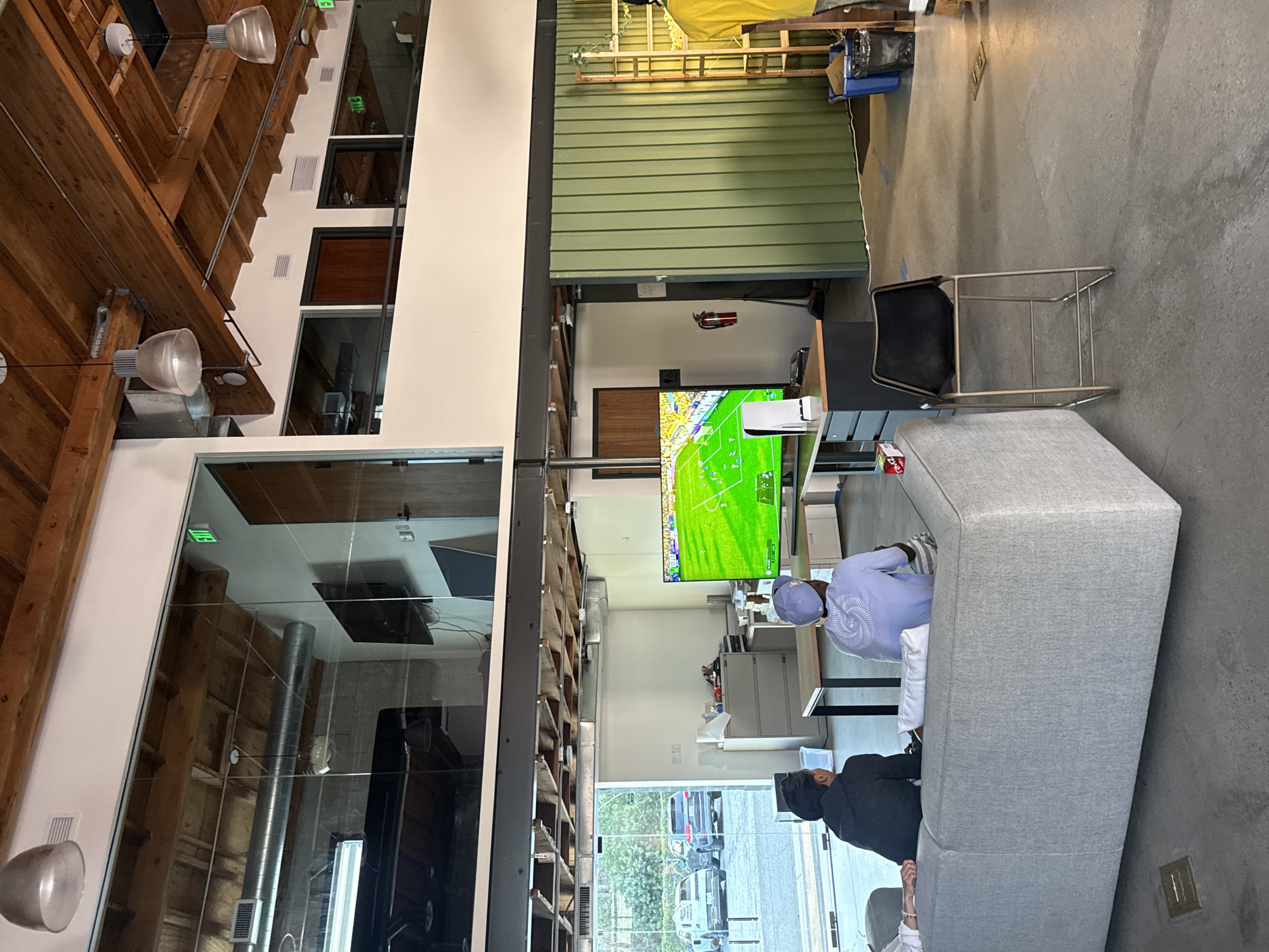 The image depicts an indoor space in what appears to be an office or collaborative workspace in Culver City, United States of America.
A TV screen displays a soccer match.
Two people are sitting on a gray couch, and one is watching the television.
Architectural details include exposed wooden beams, large windows offering a view of a city street, and industrial-style lighting fixtures. The overall environment suggests a modern, open-concept workspace.