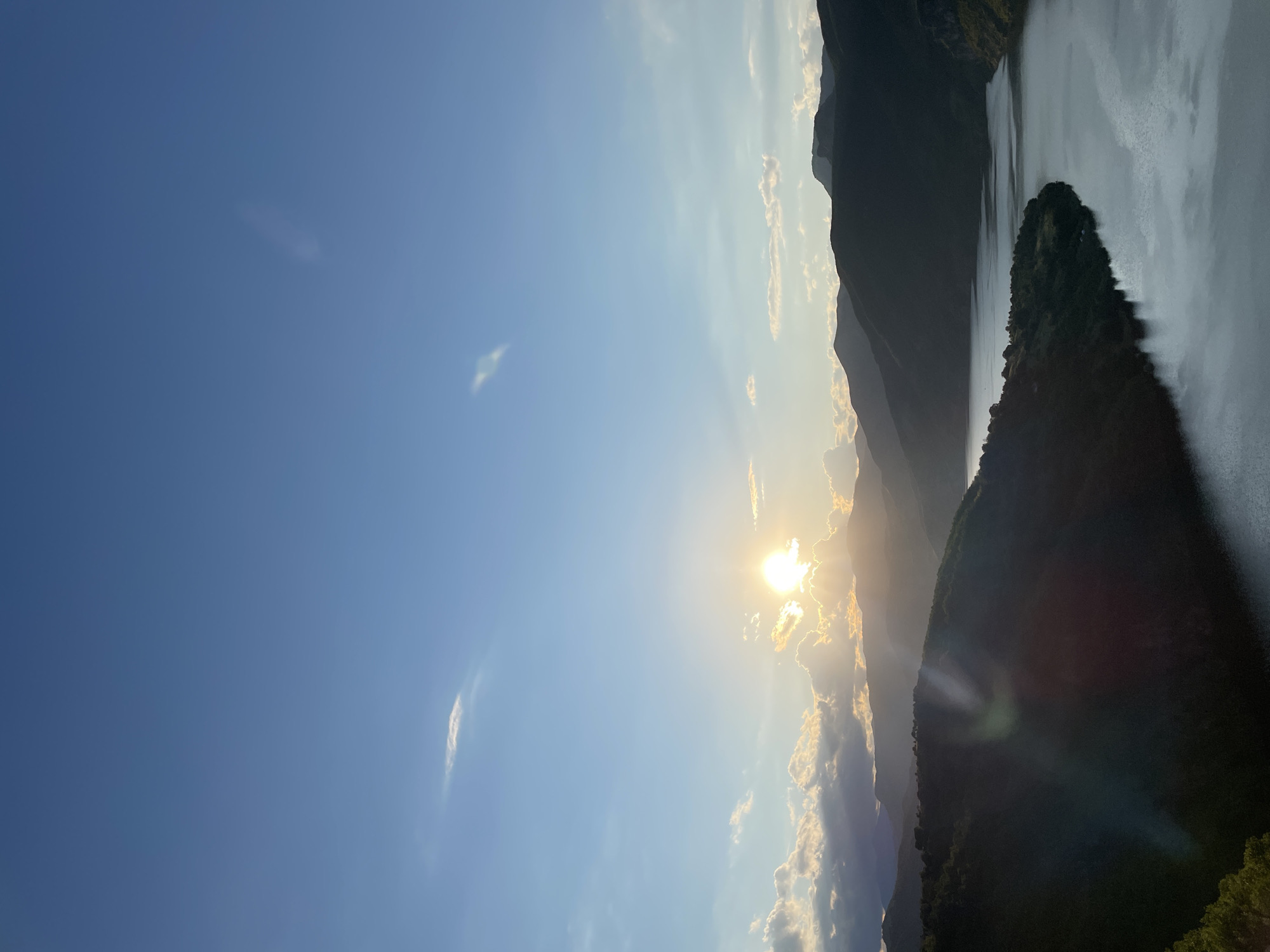 The image depicts a sunset over a mountainous landscape with a body of water in the foreground. The sun is setting behind a range of dark, silhouetted mountains, casting a warm, golden glow across the sky and illuminating a few scattered clouds. The sky is a clear blue, with some wispy clouds present. The foreground is dominated by a wide, calm body of water, likely a river or lake, reflecting the colors of the sky. Lush green vegetation covers the slopes of the mountains on either side of the water. The overall scene conveys a sense of tranquility and natural beauty. The location is identified as Borovitsa, Bulgaria. There are no people or animals visible in the image.