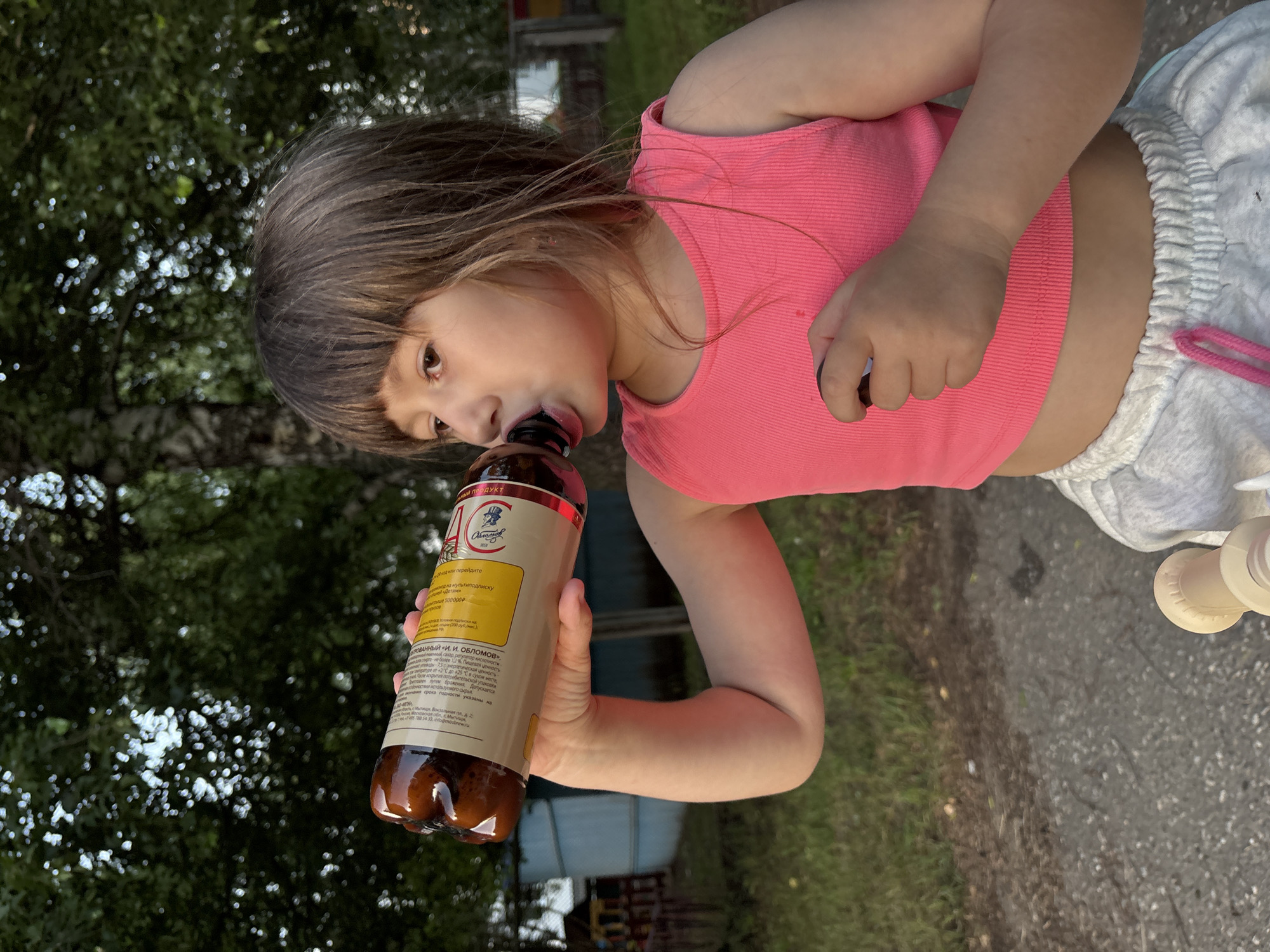 A young girl with brown hair and bangs is drinking from a brown plastic bottle. She is wearing a pink cropped tank top and grey sweatpants. The background consists of trees and a blue structure. The image appears to have been taken outdoors, possibly in a park or residential area, on a sunny day. The location is identified as Chaikovsky, Russia.