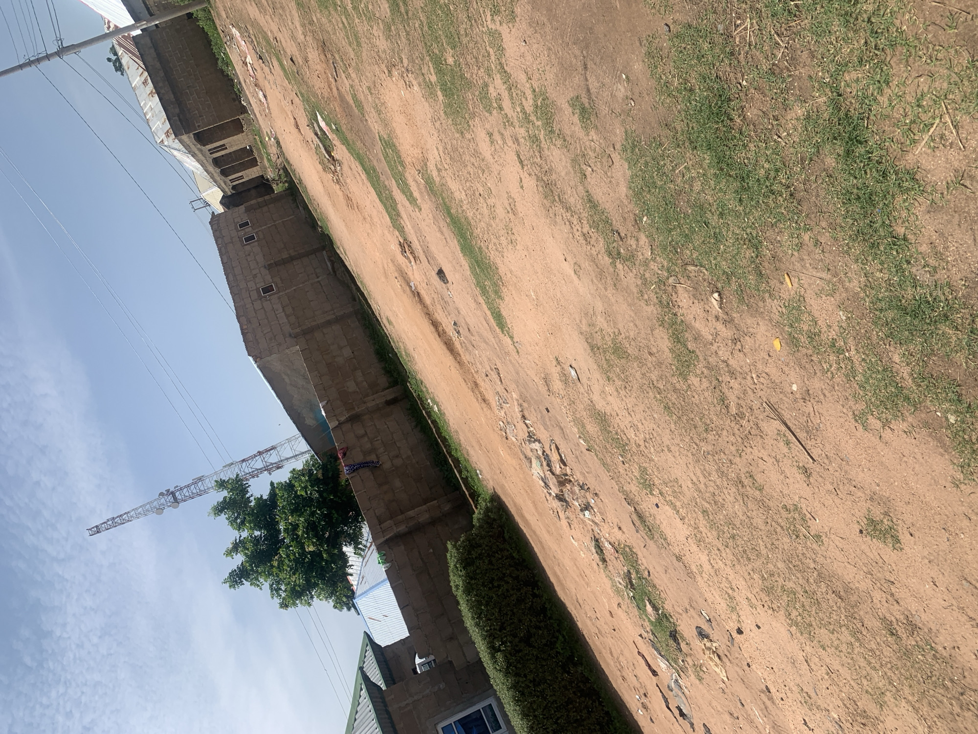 The image depicts a street scene in Jauro Yinu, Nigeria. The foreground is dominated by a reddish-brown dirt road with patches of sparse green grass. To the left, a low, neatly trimmed green hedge runs along the edge of the property. Behind the hedge, a building with a blue-framed window is visible, topped with a corrugated metal roof. A brown brick wall extends across the midground, separating the street from the residential structures beyond. Clothes are hanging on the wall.
In the background, several modest homes with corrugated metal roofs are situated. The structures appear to be constructed from beige bricks. A prominent feature in the background is a tall, red and white telecommunications tower with multiple antenna arrays. Power lines crisscross the sky above the scene. The sky is a clear, pale blue with scattered wispy clouds. The overall impression is of a rural or semi-urban environment in a hot, dry climate. There are no people visible in the image.