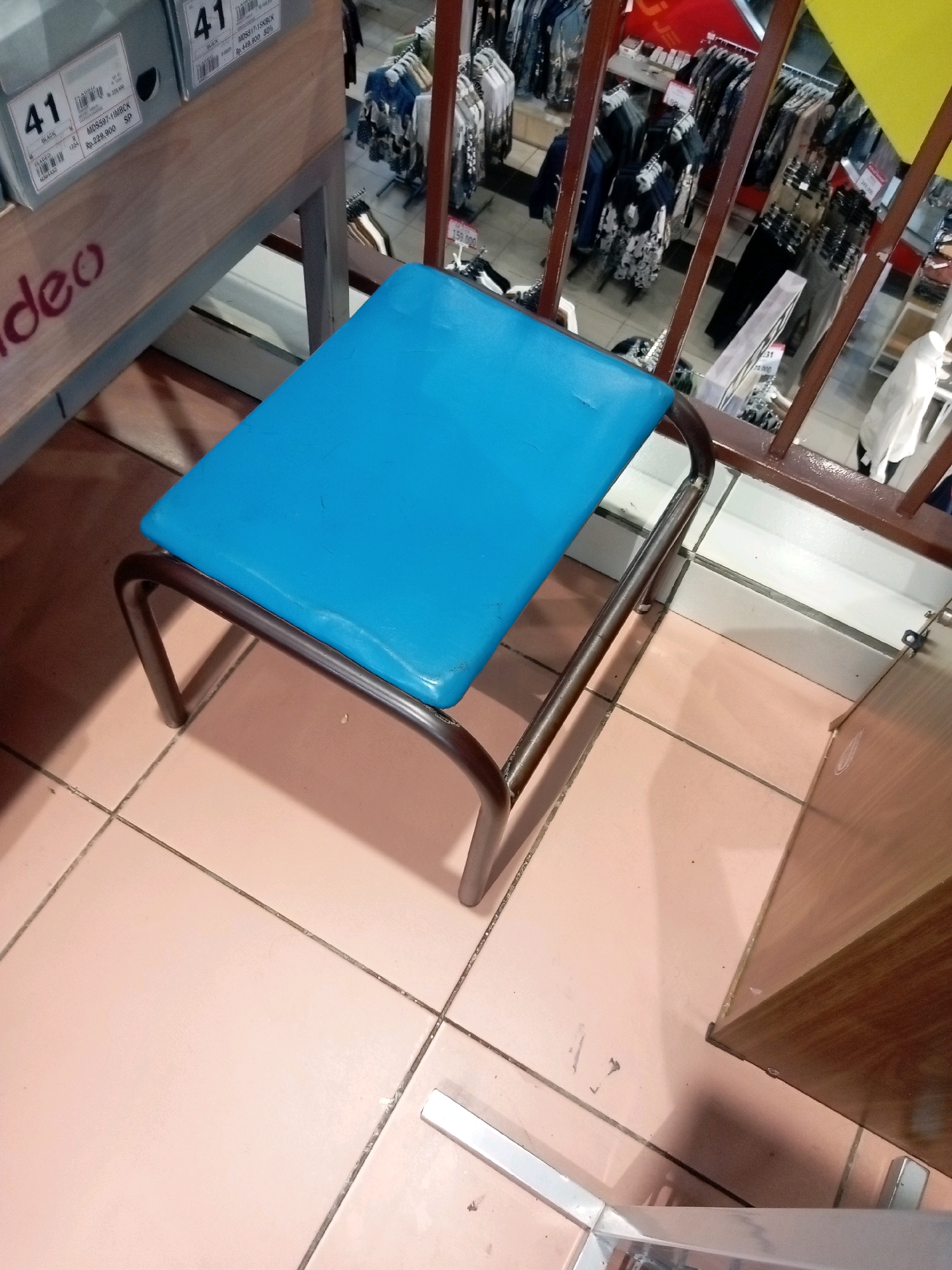 The image shows a small, blue-topped stool with a metal frame, positioned on a tiled floor. The stool is in the foreground, with its legs partially obscured by the floor tiles. Behind the stool, a store display can be seen, featuring shelves with shoe boxes and racks of clothing. The store appears to be a retail establishment, possibly a shoe or clothing store, and the context suggests it is located in Batam, Indonesia, as indicated by the shoe box labels which show Indonesian currency and text. The lighting is overhead and appears to be typical for a retail environment. There are no people visible in the image. The environment is clean but shows some scuff marks on the floor tiles and dust. The overall scene is a static, observational view of a retail space and a stool.