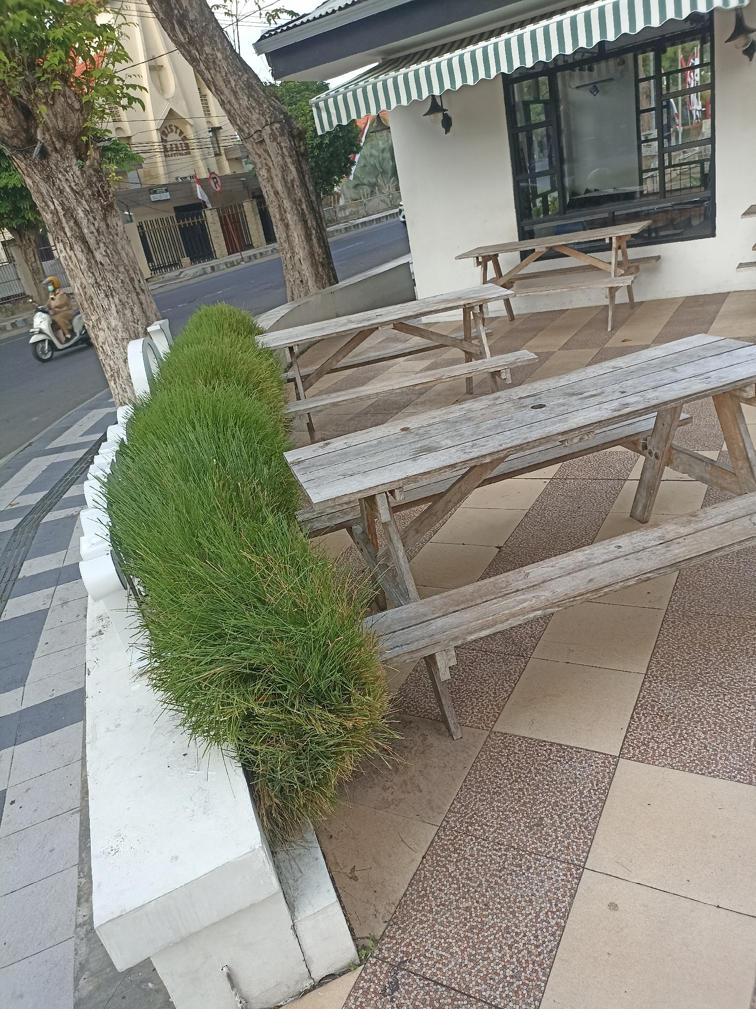 The image shows an outdoor seating area with wooden picnic tables and benches in front of a building with a striped awning. A line of lush green grass borders the seating area. In the background, a motorcycle ridden by a person in a tan uniform is visible on a street next to a building with a cross, suggesting a religious institution. The ground is paved with a mosaic of beige and speckled tiles. The overall scene depicts a tranquil urban setting.
