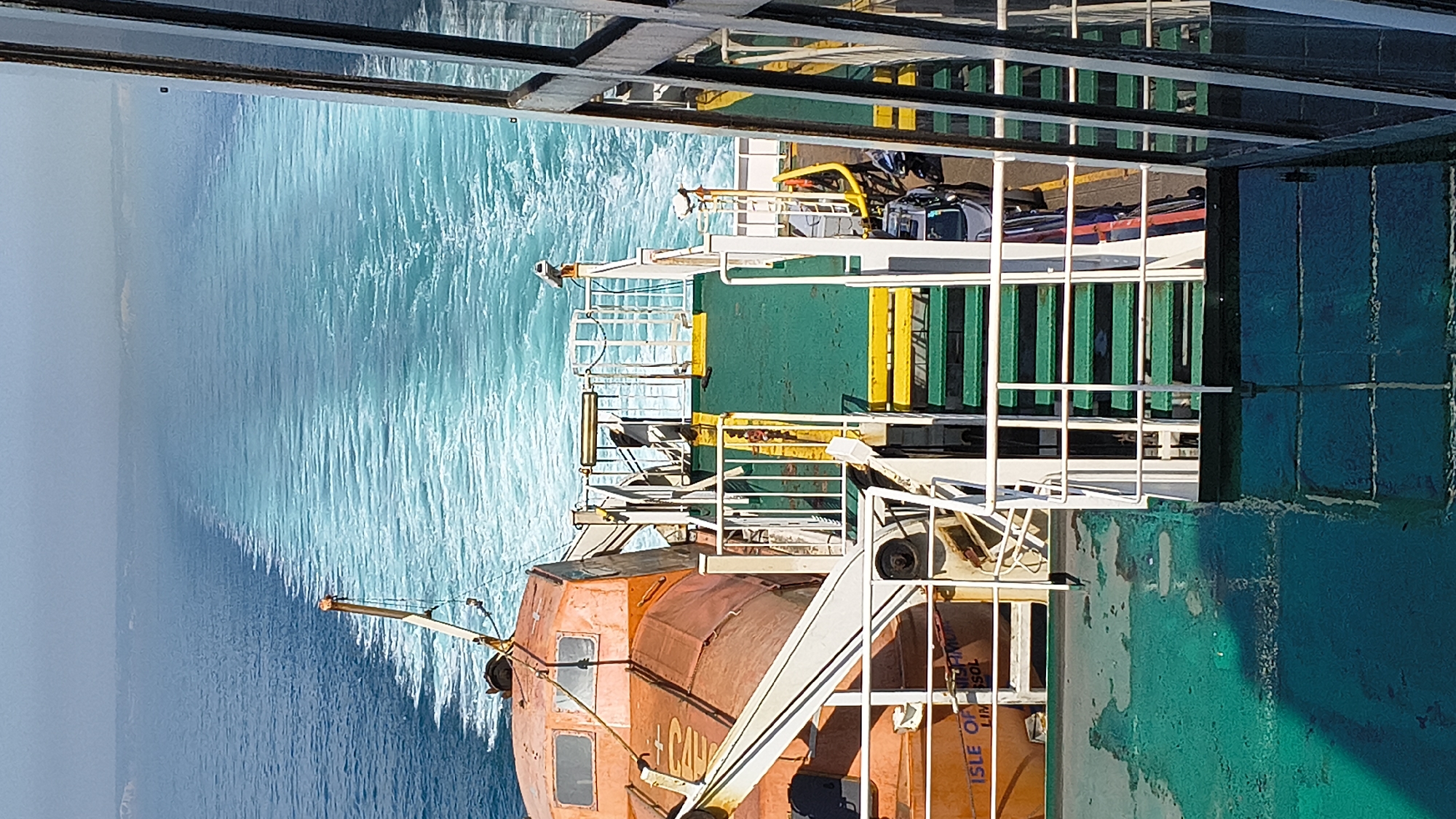 The image shows a view from a ferry deck looking out at the choppy, turquoise sea. The ferry appears to be in motion, as evidenced by the wake trailing behind it. On the deck, various objects are visible, including a bright orange lifeboat, yellow and green barriers, and what appears to be a vehicle secured with straps. White railings and structures of the ferry's upper decks are also prominent.
The lighting suggests it is daytime, with bright sunlight reflecting off the water and the ferry's surfaces. The sea conditions appear to be moderately rough, with whitecaps indicating wind and waves.
Partial text is visible on the side of the orange lifeboat, reading "+ CAV" and "ISLE OF LIM...SOL". The lifeboat is also marked with "ISLE OF MAN SHIPOWNERS" and "LIMERICK" with "LIM" partially obscured. This suggests the lifeboat is associated with a vessel from the Isle of Man.
The overall scene is one of transit and maritime activity. The perspective is from a higher deck of the ferry, looking down and across. No people are clearly visible in the frame. The scene is functional and industrial, typical of a working ferry, but the vibrant blue of the sea adds a natural beauty.