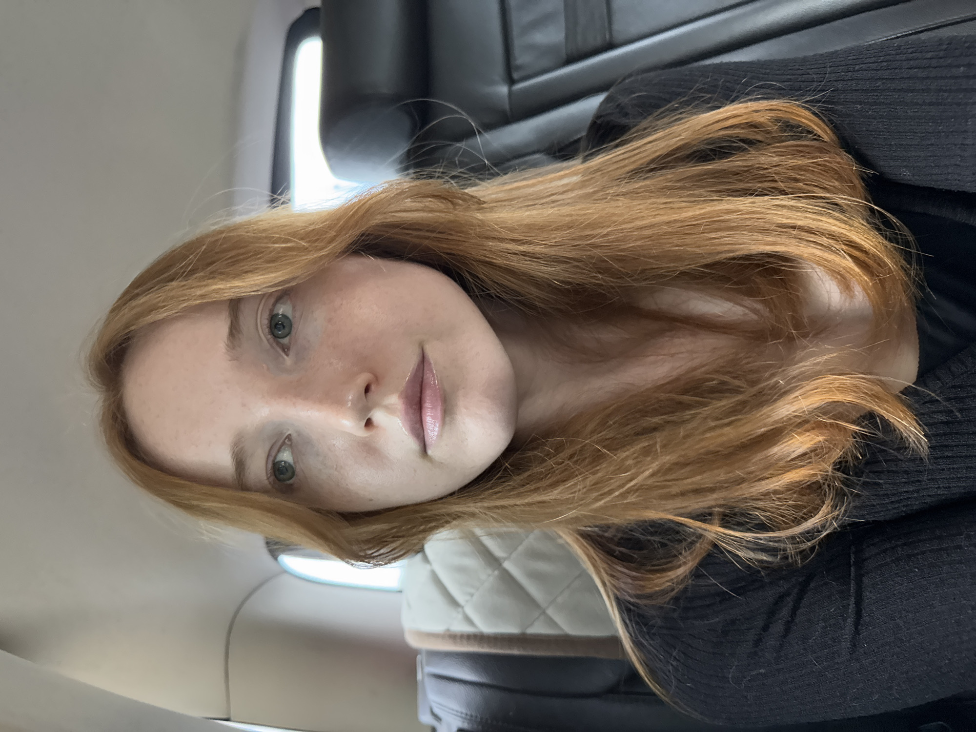 A fair-skinned person with red hair and green eyes is seated in a vehicle. The person is wearing a black top. The background shows elements of a vehicle interior, including a headrest with a quilted pattern and dark leather seats. The location is not possible to deduce from this image.