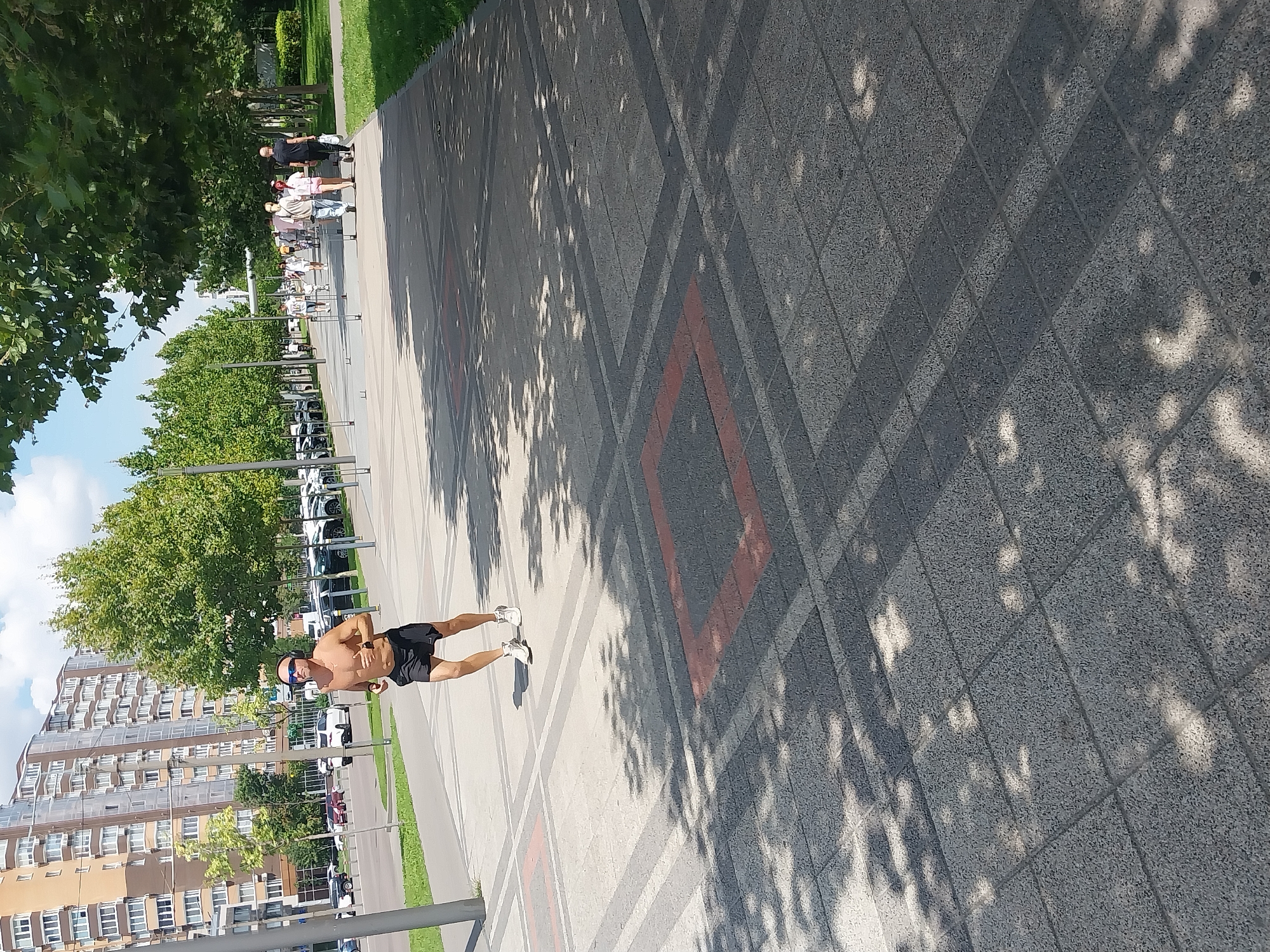 In Dnipro, Ukraine, on a bright sunny day, a man is jogging on a paved pedestrian walkway. He is shirtless, wearing black athletic shorts, white sneakers, and dark sunglasses. He appears to be wearing headphones, suggesting he is listening to music or a podcast while exercising. The walkway is wide and made of patterned grey paving stones with red rectangular accents. Trees with full green foliage line the walkway on the right, casting dappled shadows across the ground. In the background, on the left side of the image, is a modern, multi-story residential building with many windows. Further down the walkway, other pedestrians are visible, walking in various directions. To the right of the walkway is a strip of green grass. Cars are parked along the street to the left of the building. The overall scene depicts an urban park or promenade area on a pleasant day.