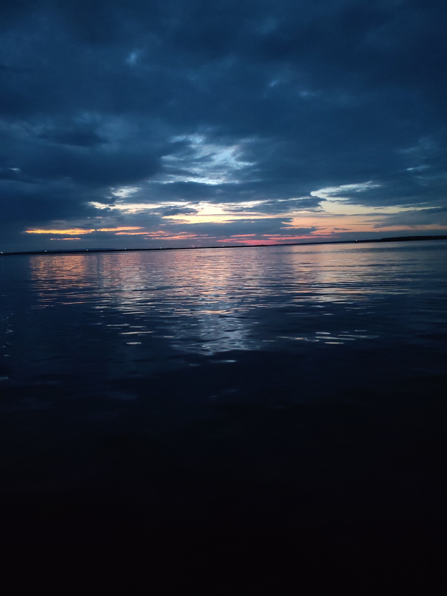 The image depicts a wide body of water, likely a reservoir or river, at dusk. Dark, brooding clouds dominate the upper portion of the frame, with hints of orange and pink light breaking through the horizon. The water's surface is rippled, reflecting the muted colors of the sky. In the distance, along the far shore, faint lights are visible, suggesting a populated area. The foreground of the image fades into darkness, implying the viewer is positioned at the water's edge or on a vessel close to the shore. The overall atmosphere is serene and slightly melancholic, characteristic of twilight on a large body of water. This scene is consistent with views of the Kama River or the Votkinskoye Reservoir near the city of Chaikovsky, Russia.