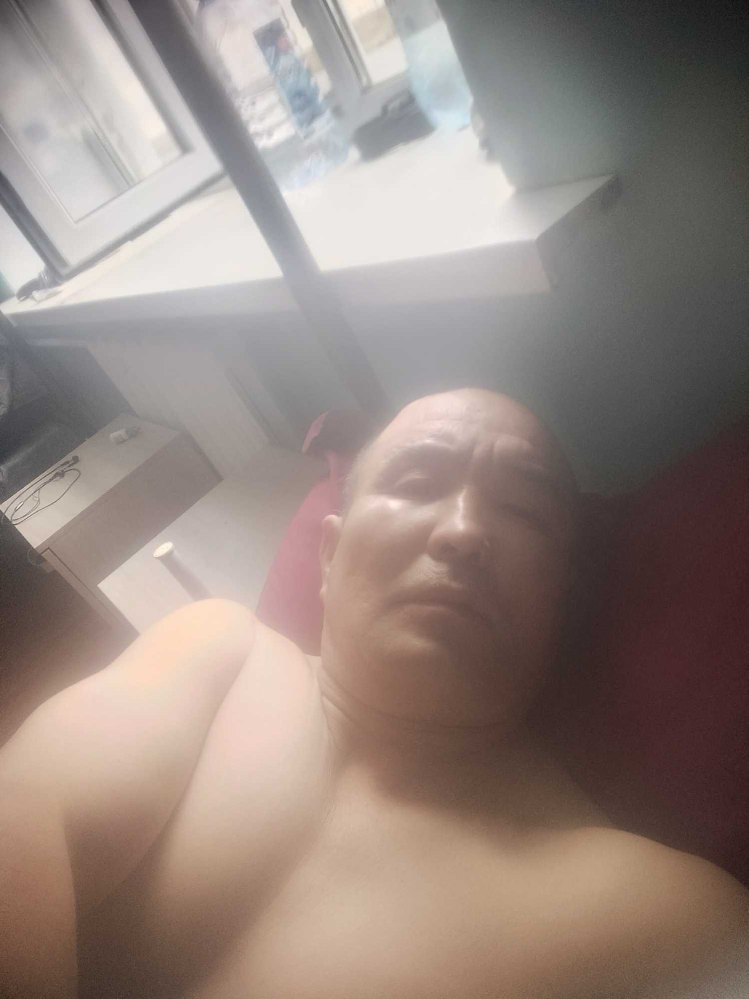 An individual is lying down, facing the camera, in what appears to be a bedroom. The person is bald and has a fair complexion. They are shirtless, with their left shoulder and arm prominently visible in the foreground. The background suggests an indoor setting with a window and furniture, including what looks like a dresser or desk with some items on it. The overall lighting is soft, possibly from natural light coming through the window. The image is taken at a close range, focusing on the person's upper body and face. There are no discernible features that would indicate the location is specifically in Jezqazğan, Kazakhstan.