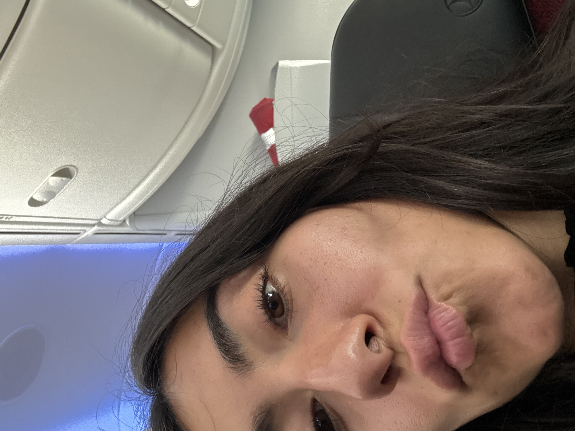 A woman with long dark hair is taking a selfie on an airplane. The background includes overhead cabin lights and a monitor screen. The location is likely in the air, but the airplane details suggest a commercial flight, not necessarily limited to Mexico City, Mexico.