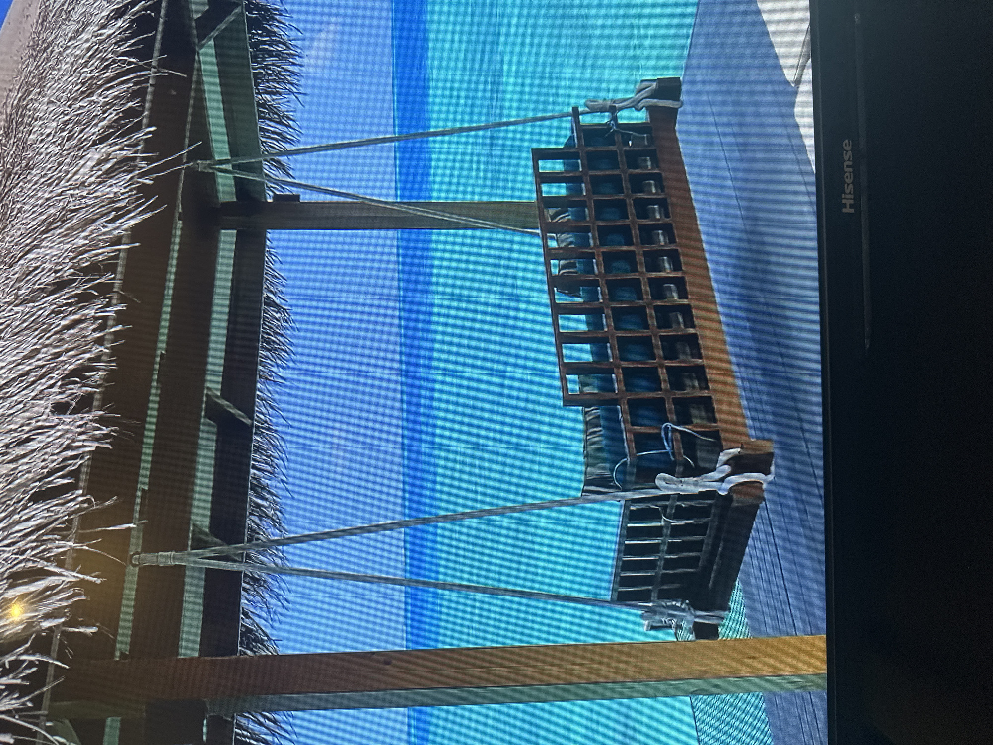 The image shows a wooden swing with cushions hanging from a thatched roof structure overlooking a bright turquoise ocean. The swing is suspended by ropes from a beam of the structure. The structure has wooden posts and a slatted wooden floor. The ocean is calm with a clear horizon. The scene is brightly lit, suggesting a sunny day. There are no people or animals visible in the image. The Hisense logo is visible at the bottom of the image. The location is not in Kyiv, Ukraine; it appears to be a tropical or subtropical resort.